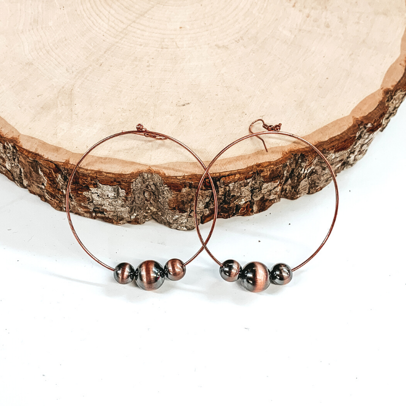 Copper open circle drop earrings with copper beads in varying sizes at the bottom of the earrings. These earrings are pictured laying against a piece of wood. 