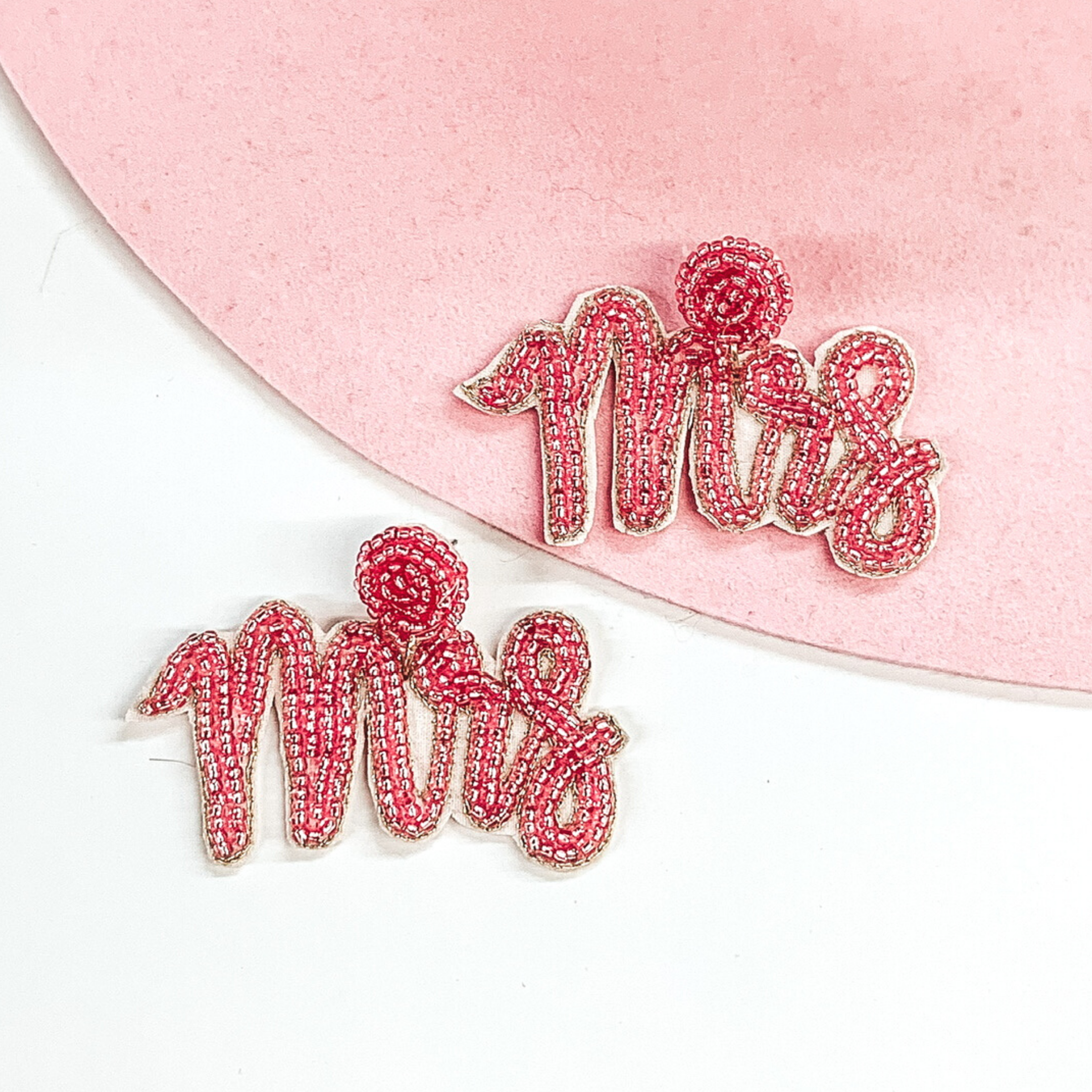 Pink beaded circle post back studs with a hanging pendant beaded "Mrs" in cursive. These earrings are pictured on a white and light pink background. 