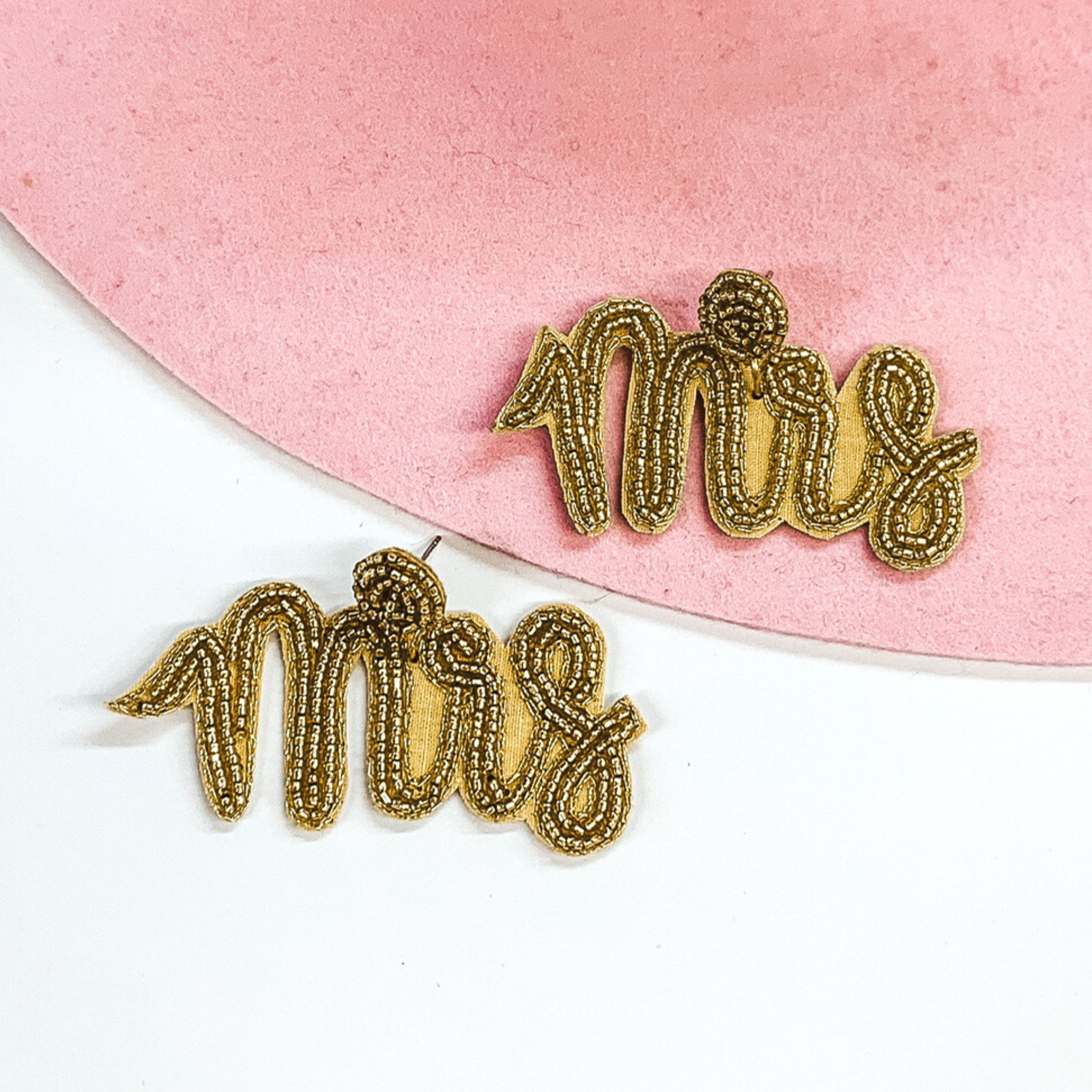 Gold beaded circle post back studs with a hanging pendant beaded "Mrs" in cursive. These earrings are pictured on a white and light pink background. 