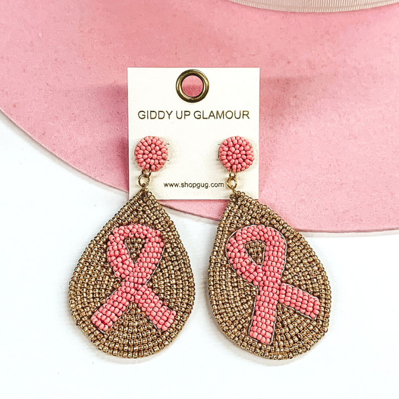 Light pink beaded circle studs with hanging beaded teardrop pendant. The pendant has a gold beaded background with a center beaded light pink ribbon These earrings are pictured on a white and pink background. 
