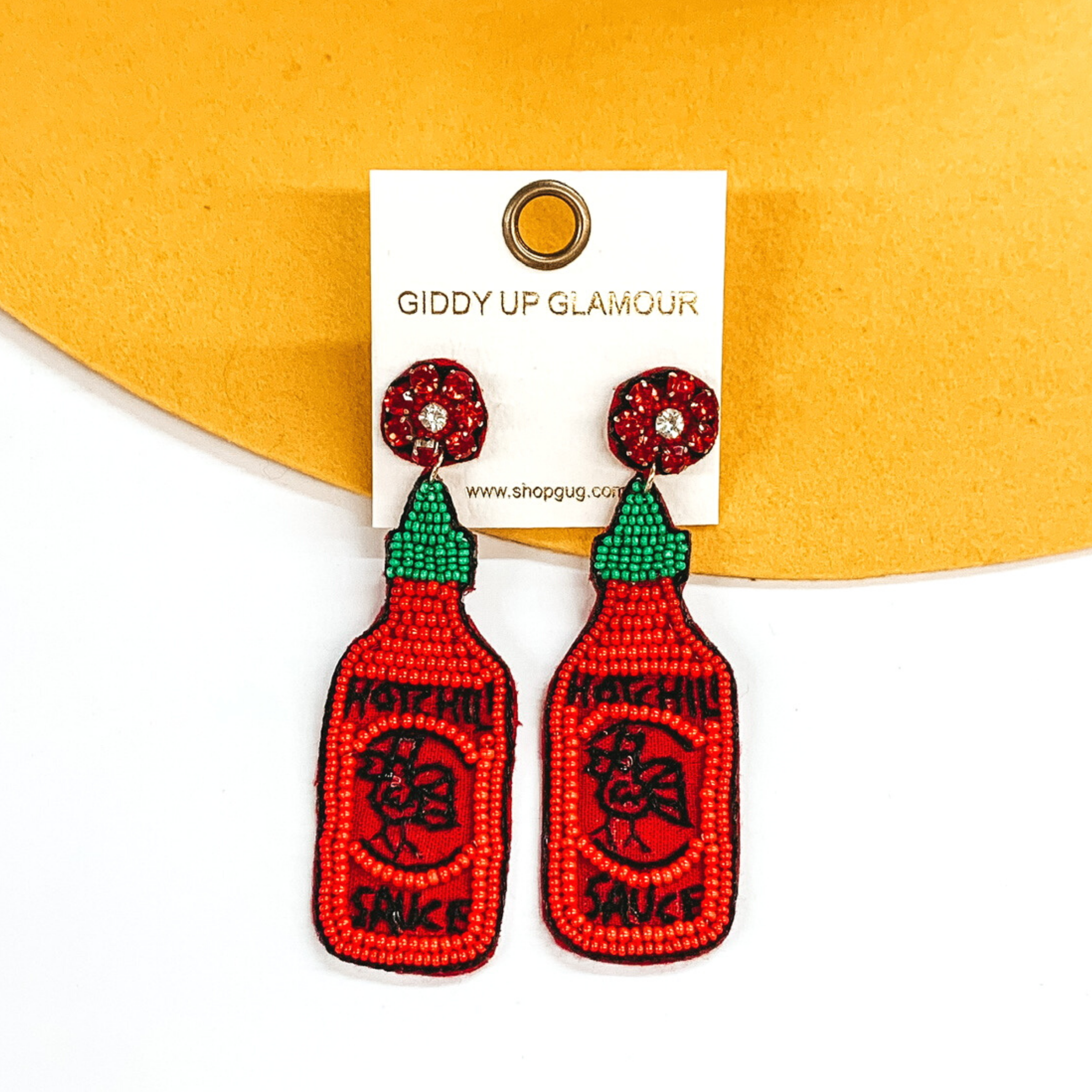 Red crystal beaded circle studs with hanging beaded bottle pendant. The pendant has a green beaded cap and red beaded bottle. The center of the bottle has a black stitched design. These earrings are pictured on a white and yellow background. 