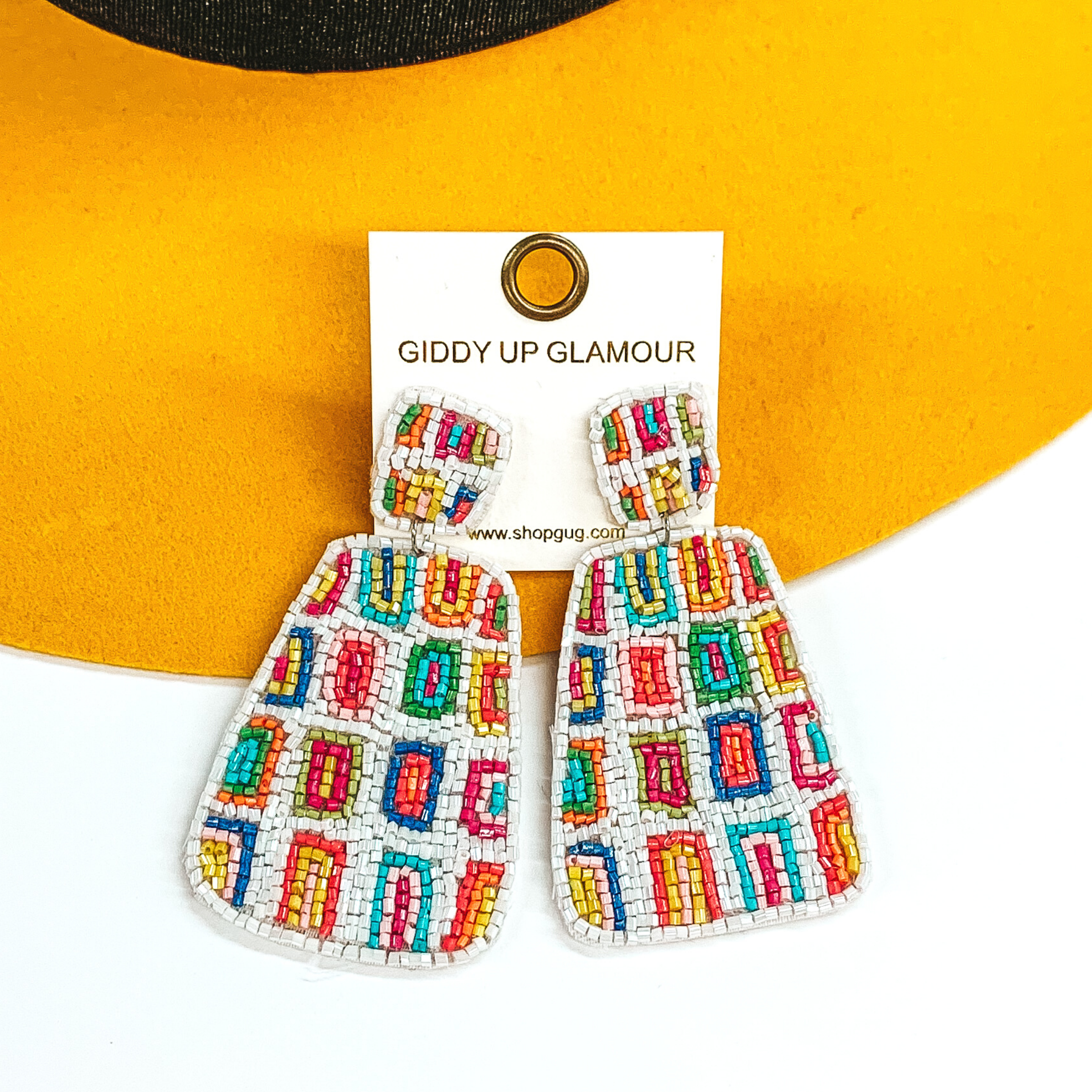 Irregular square shapes studs with an irregular shaped rectangle hanging pendant. Both has a white beaded background with multicolored rectangles throughout. These earrings are pictured on a white and yellow background. 