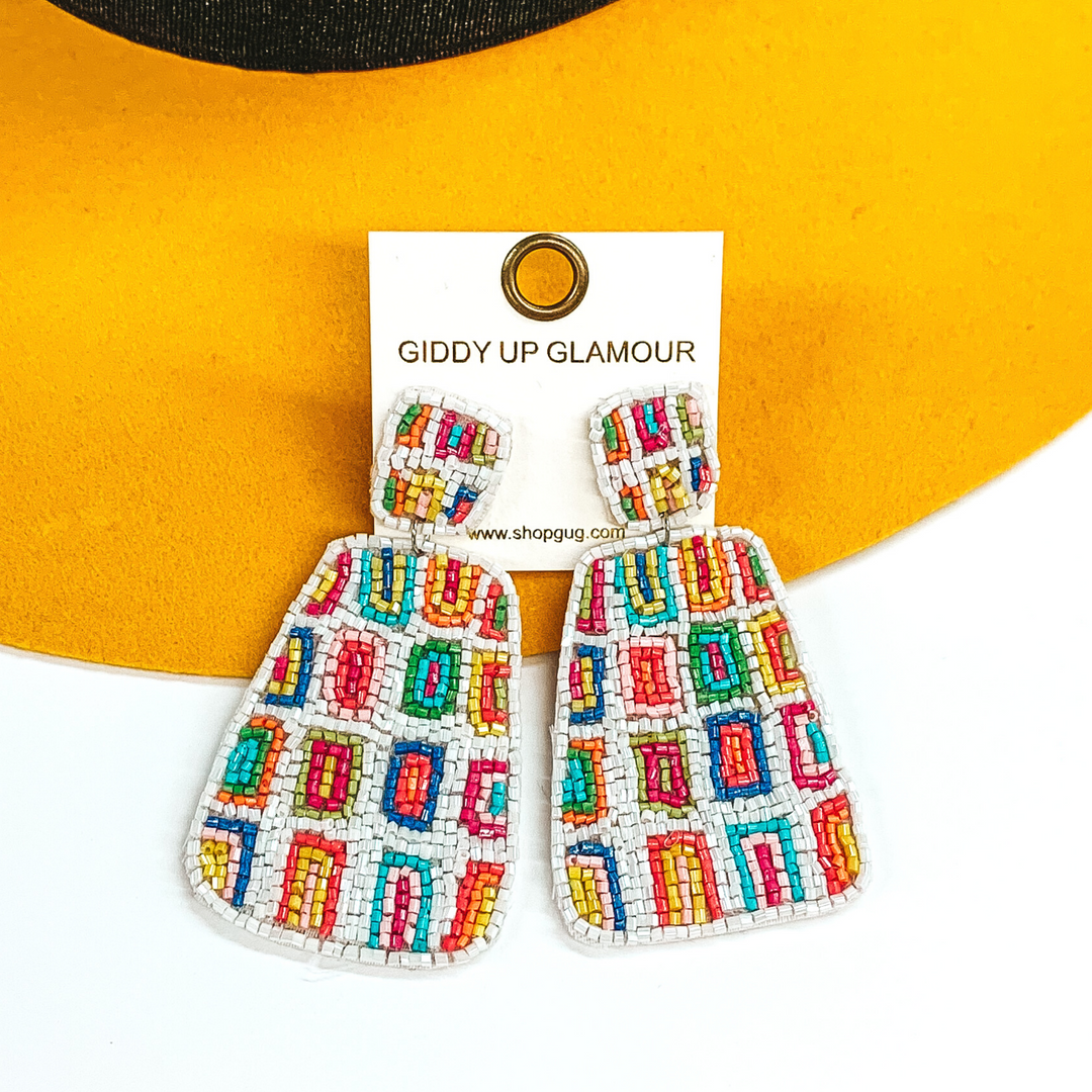 Irregular square shapes studs with an irregular shaped rectangle hanging pendant. Both has a white beaded background with multicolored rectangles throughout. These earrings are pictured on a white and yellow background. 