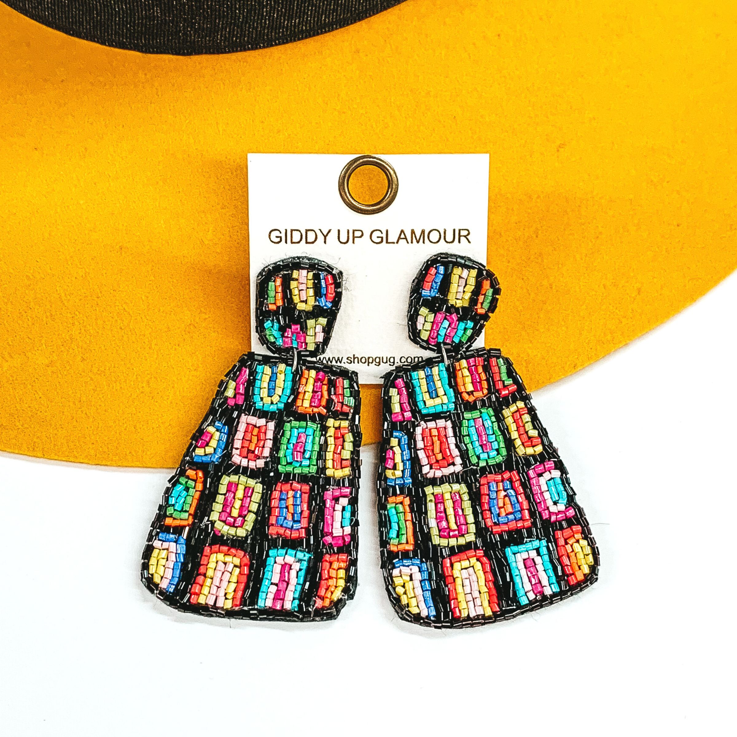 Irregular square shapes studs with an irregular shaped rectangle hanging pendant. Both has a black beaded background with multicolored rectangles throughout. These earrings are pictured on a white and yellow background. 