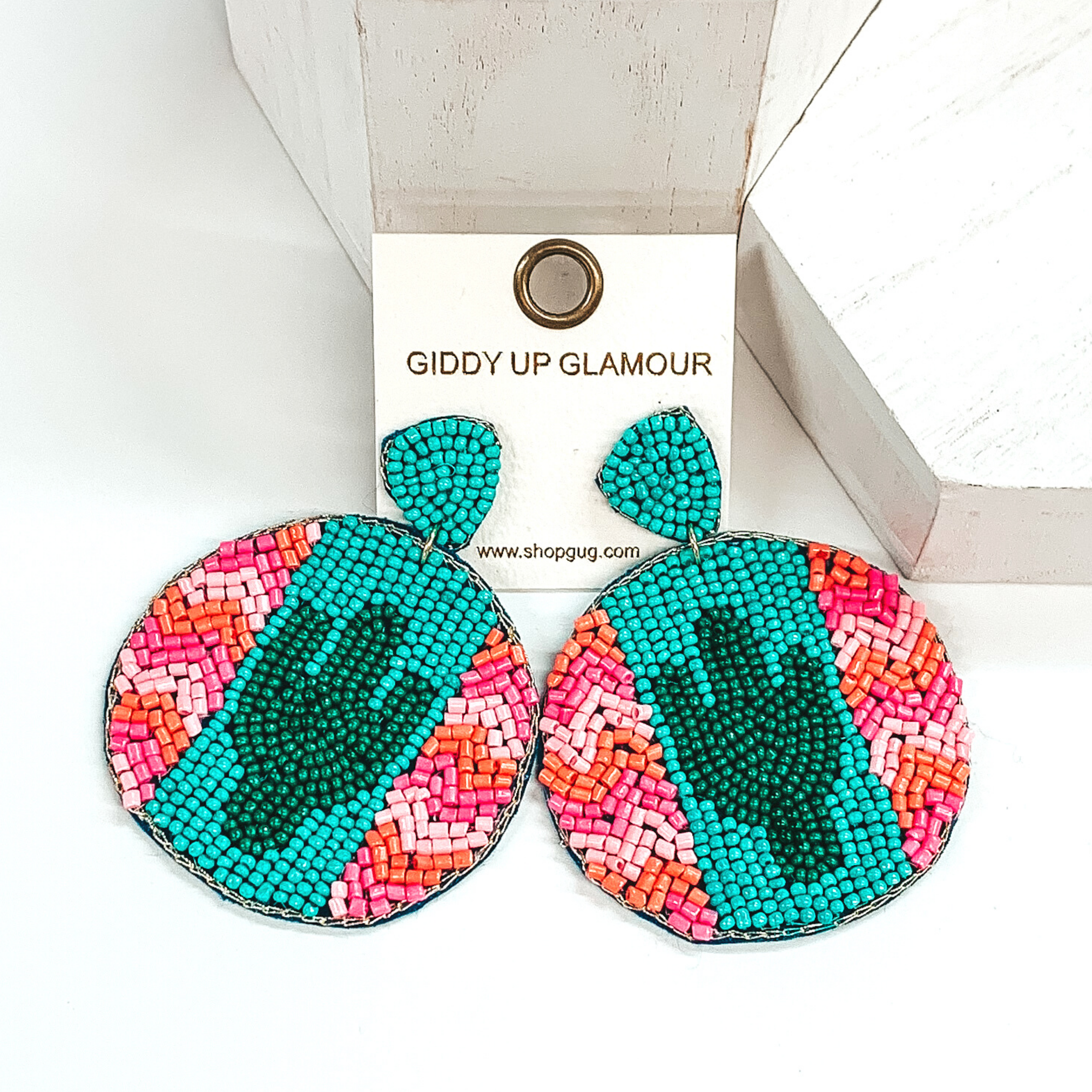 Turquoise beaded triangle studs with a beaded hanging circle. The circle has a turquoise colored stripe down the middle with a dark green cactus in the center. On the sides of the stripes these is a light pink, pink, and orange chevron pattern. These earrings are pictured on a white background with white blocks at the top of the picture. 
