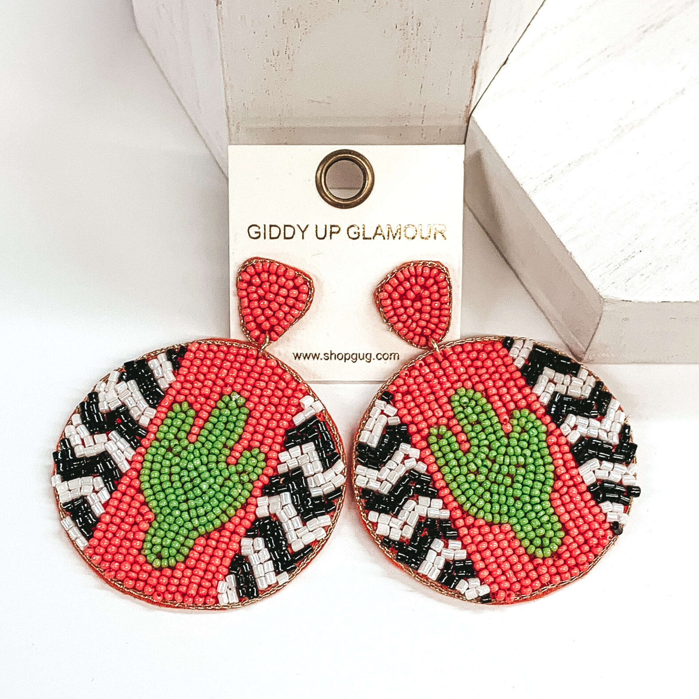 Coral beaded triangle studs with a beaded hanging circle. The circle has a coral colored stripe down the middle with a green cactus in the center. On the sides of the stripes these is a black and white chevron pattern. These earrings are pictured on a white background with white blocks at the top of the picture. 