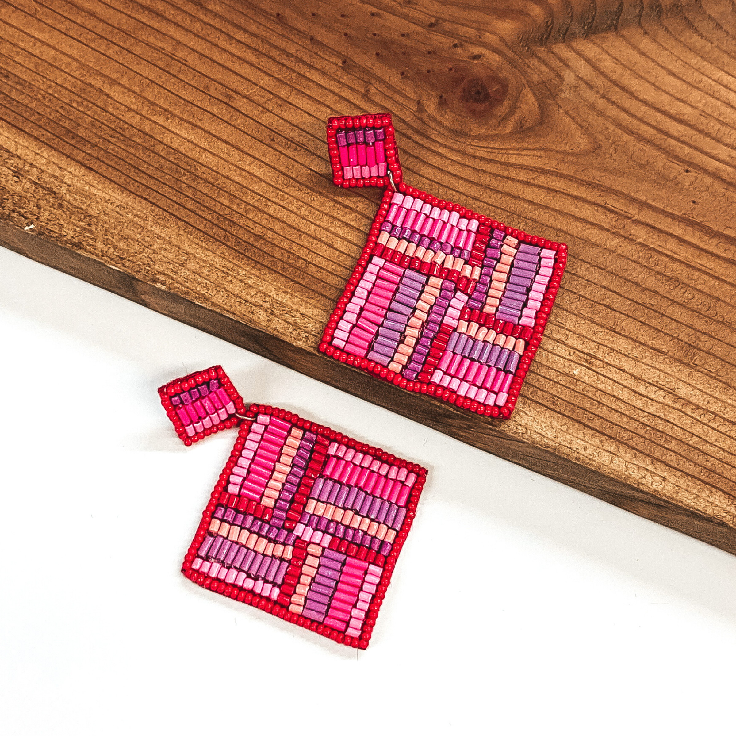 Square beaded studs with a beaded hanging square. Both are outlined in red and include a light pink, dark pink, purple, nude, and red beaded design. These earrings are pictured with one earrings on a white background and one on a dark piece of wood. 