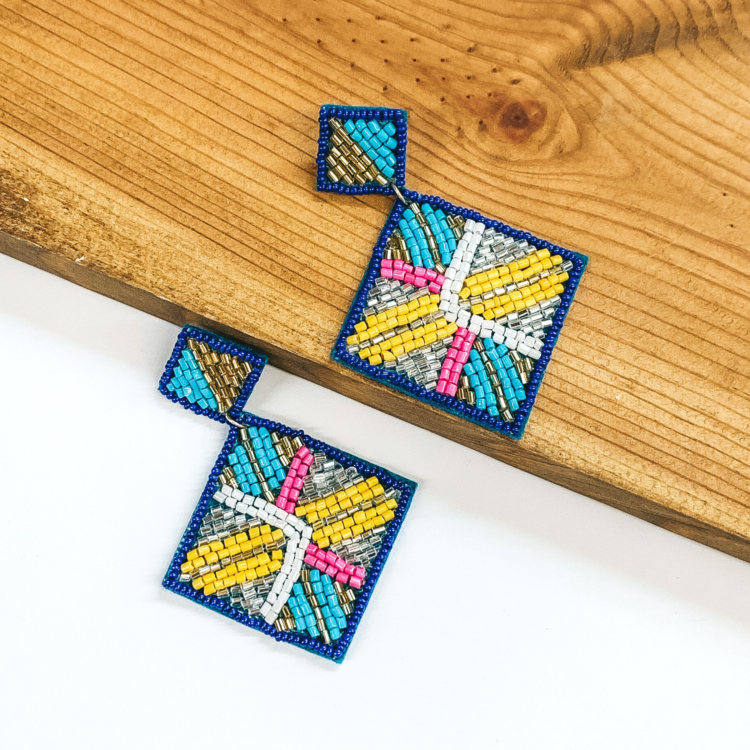 Square beaded studs with a beaded hanging square. Both are outlined in blue and include a light pink, white, yellow, light blue, gold, and silver beaded design. These earrings are pictured with one earrings on a white background and one on a dark piece of wood. 