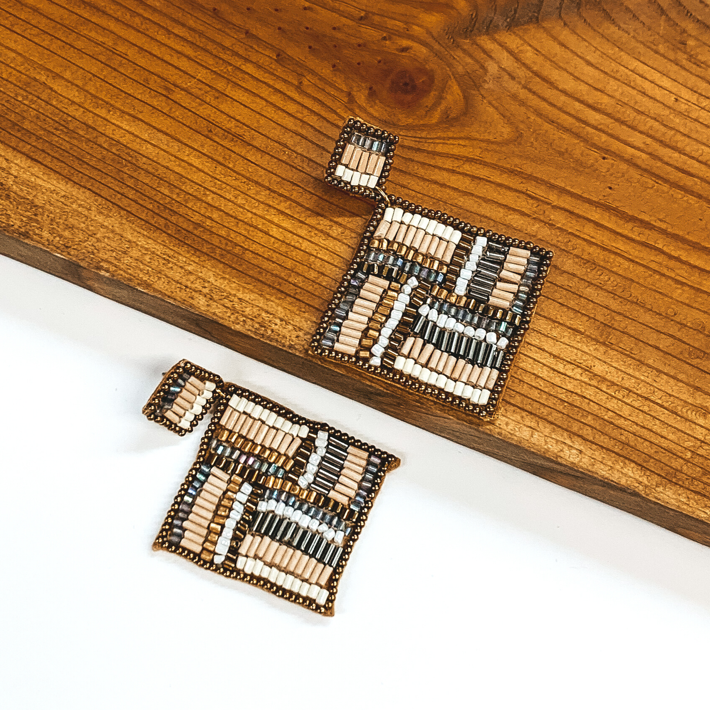 Square beaded studs with a beaded hanging square. Both are outlines in bronze and include a gold, cream, grey, bronze, and nude beaded design. These earrings are pictured with one earrings on a white background and one on a dark piece of wood. 