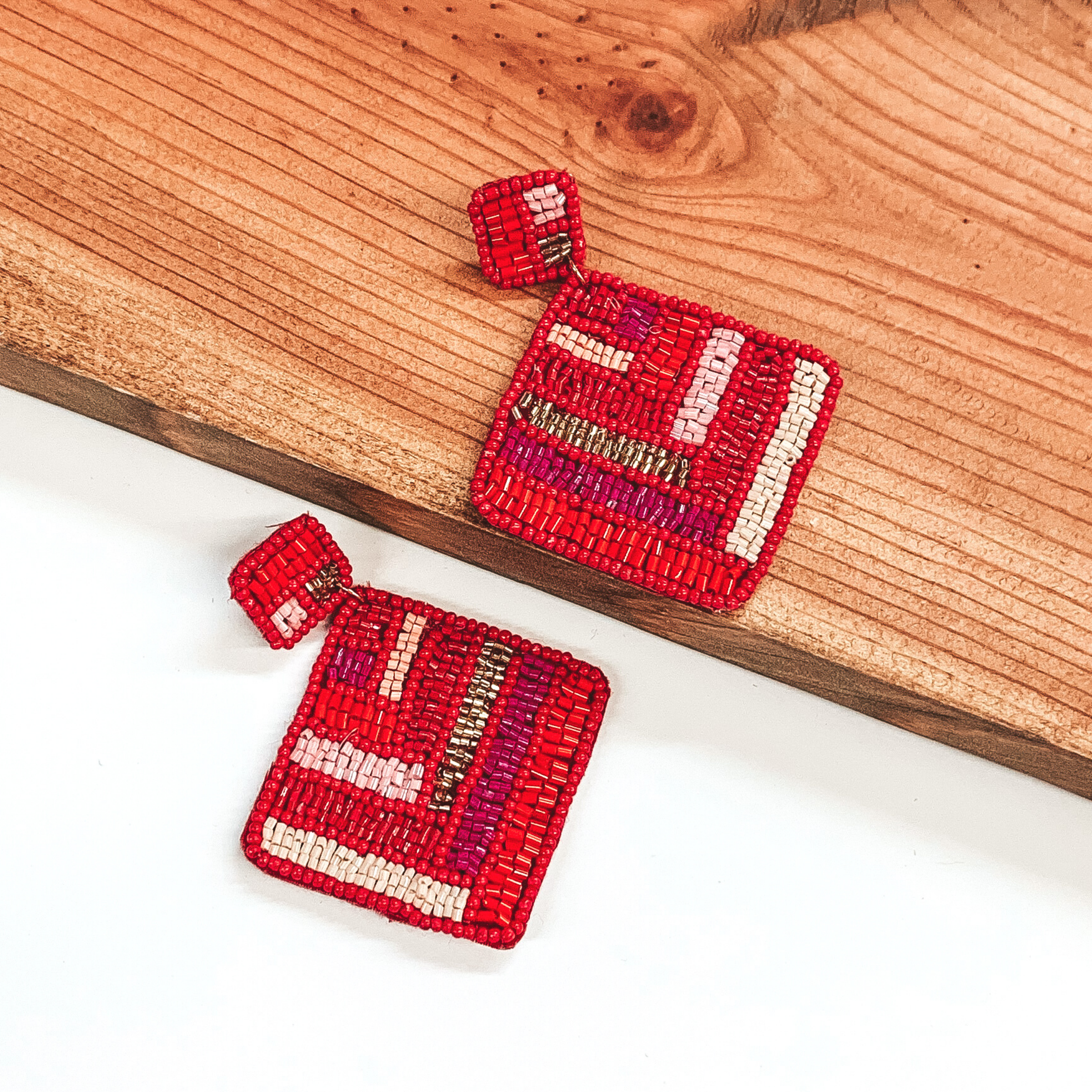 Square beaded studs with a beaded hanging square. Both are outlined in red and include a light pink, dark purple, nude, gold, and red chevron beaded design. These earrings are pictured with one earrings on a white background and one on a dark piece of wood. 