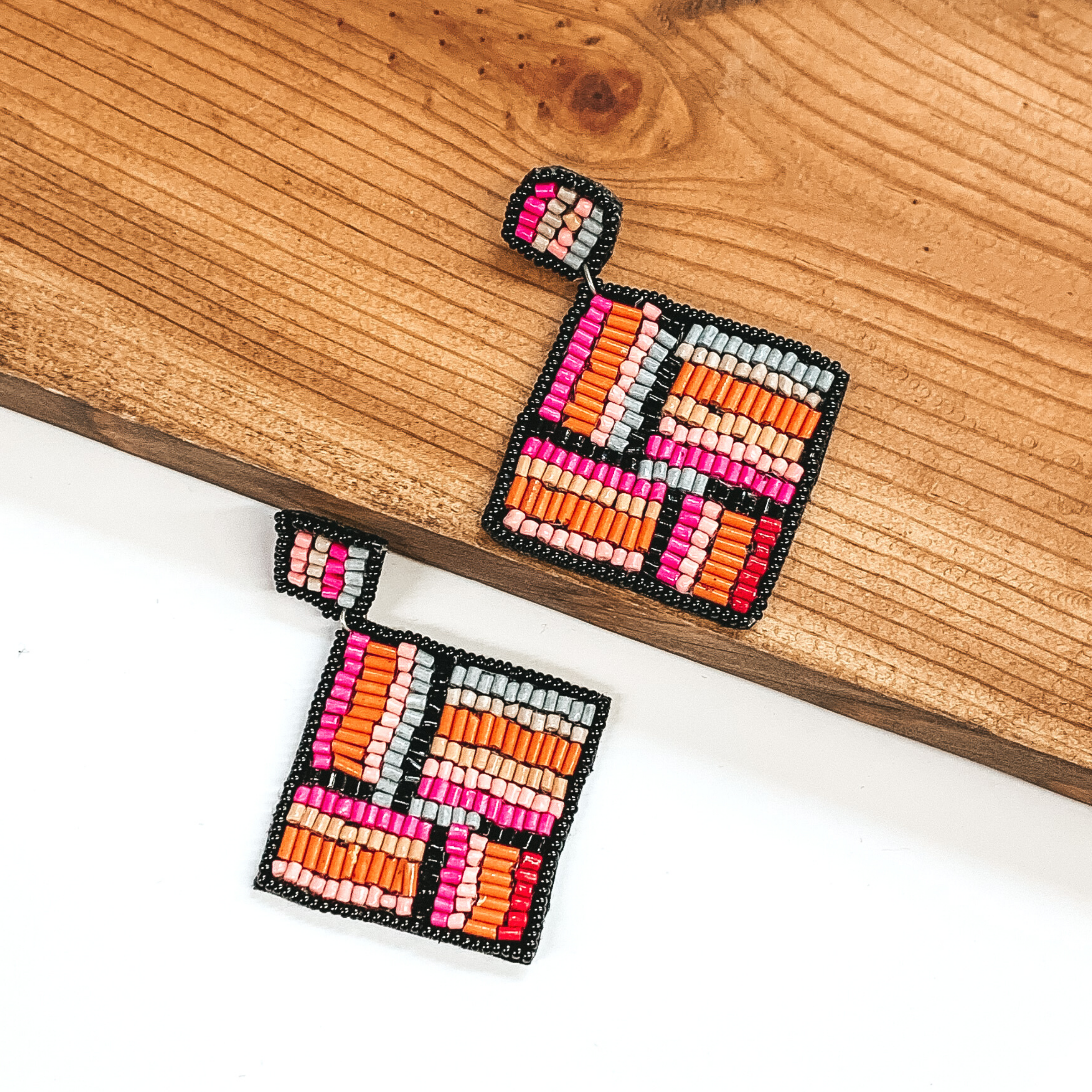 Square beaded studs with a beaded hanging square. Both are outline in black and include a pink, light pink, orange, grey, red, and nude beaded design. These earrings are pictured with one earrings on a white background and one on a dark piece of wood. 