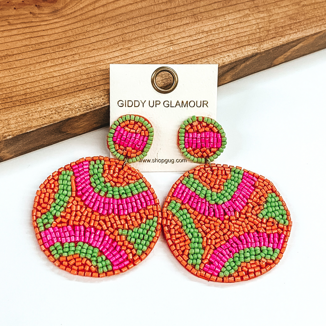 Circle beaded studs with a beaded hanging circle. Both include a orange, pink, and lime green design. These earrings are pictured in front of dark colored wood on a white background. 