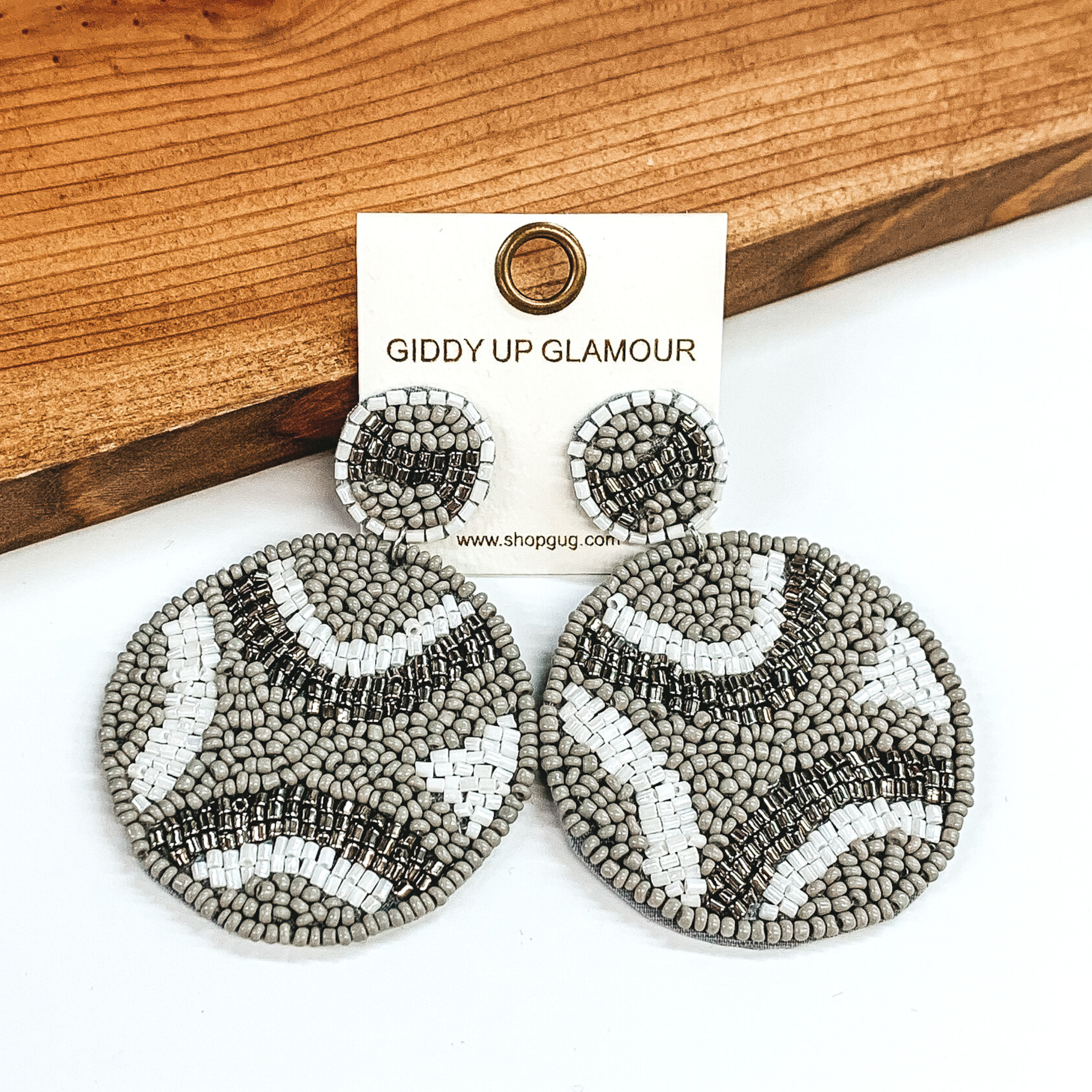 Circle beaded studs with a beaded hanging circle. Both include a white, grey, and shiny grey design. These earrings are pictured in front of dark colored wood on a white background. 
