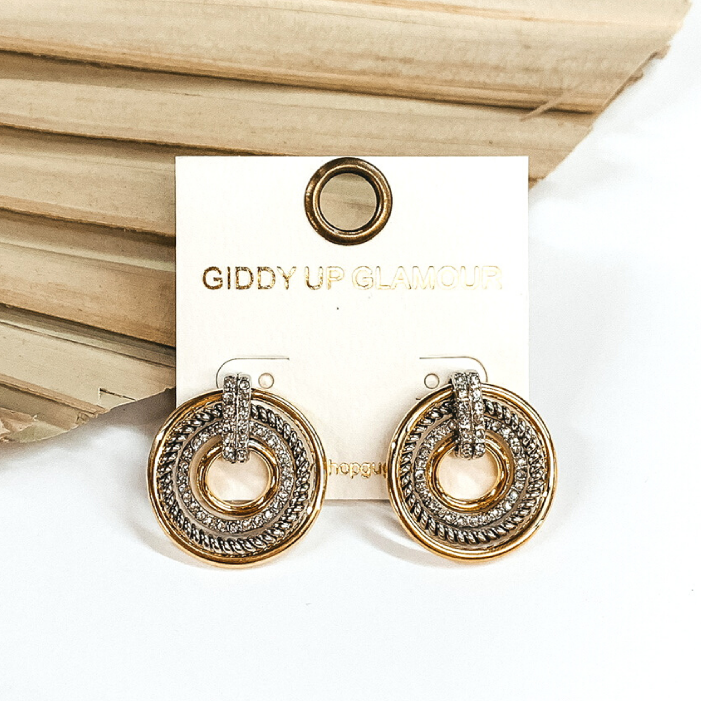 These earrings include gold rings, a silver rope ring, and a clear crystal ring connected together with a clear crystal connecter. These earrings are pictured in front of a dried sage green palm leaf on a white background. 