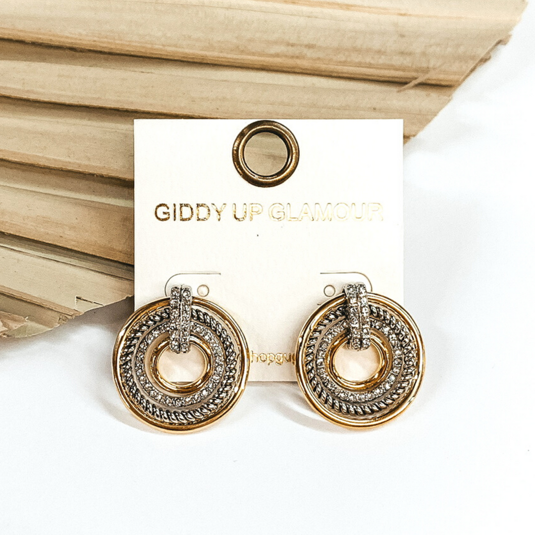 These earrings include gold rings, a silver rope ring, and a clear crystal ring connected together with a clear crystal connecter. These earrings are pictured in front of a dried sage green palm leaf on a white background. 