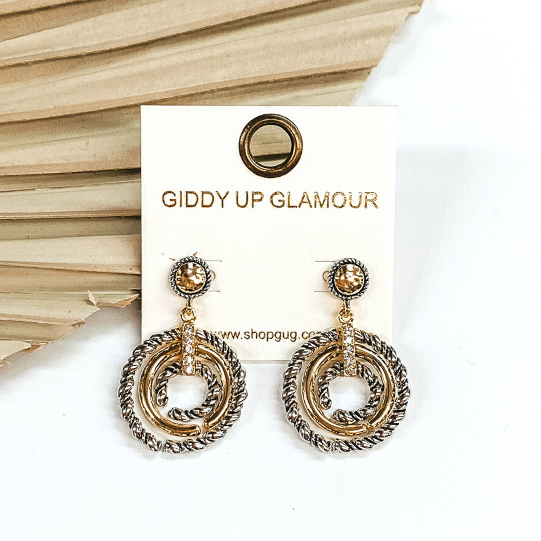 Gold studs with silver rope outline with a hanging circle pendant. The circle pendant includes silver rope rings and a gold ring. These rings are connected with a gold and clear crystal connector. These earrings are pictured on front of safe green dried palm leaf on a white background. 