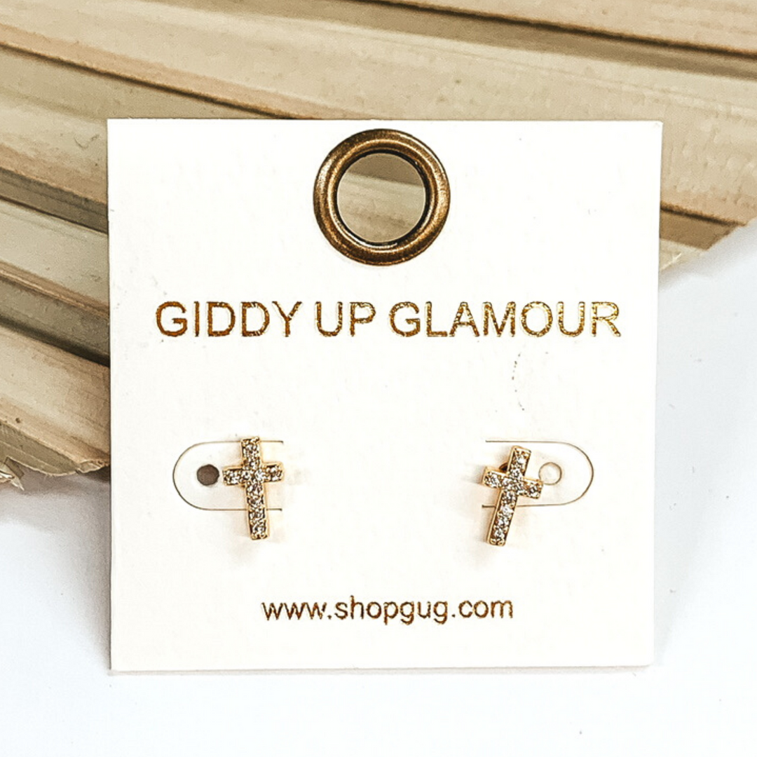 Small, gold cross stud earrings with clear crystals. These earrings are pictured on a white earrings card on a white and brown background.