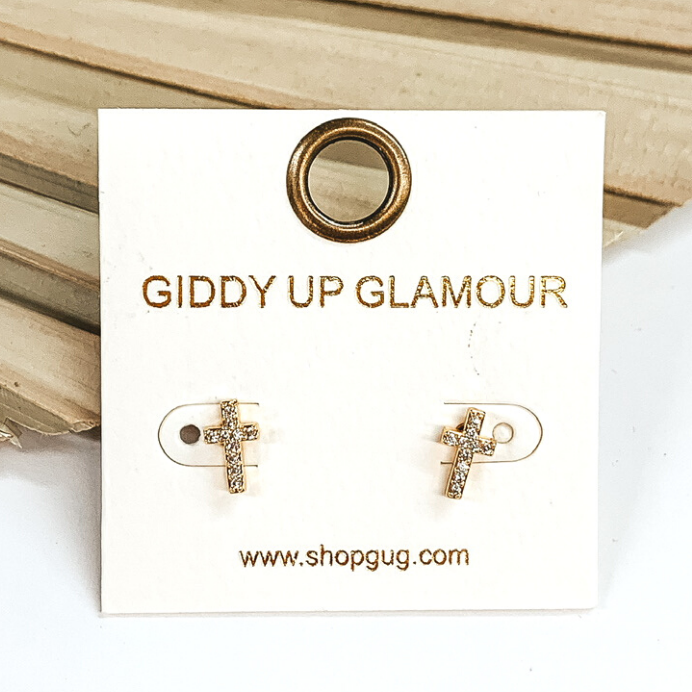 Small, gold cross stud earrings with clear crystals. These earrings are pictured on a white earrings card on a white and brown background.