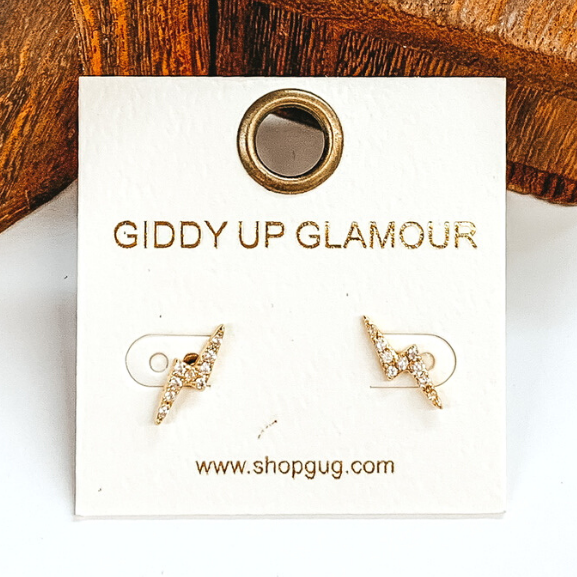 Small, gold thunder bolt stud earrings with clear crystals. These earrings are pictured on a white earrings card on a white and brown background.