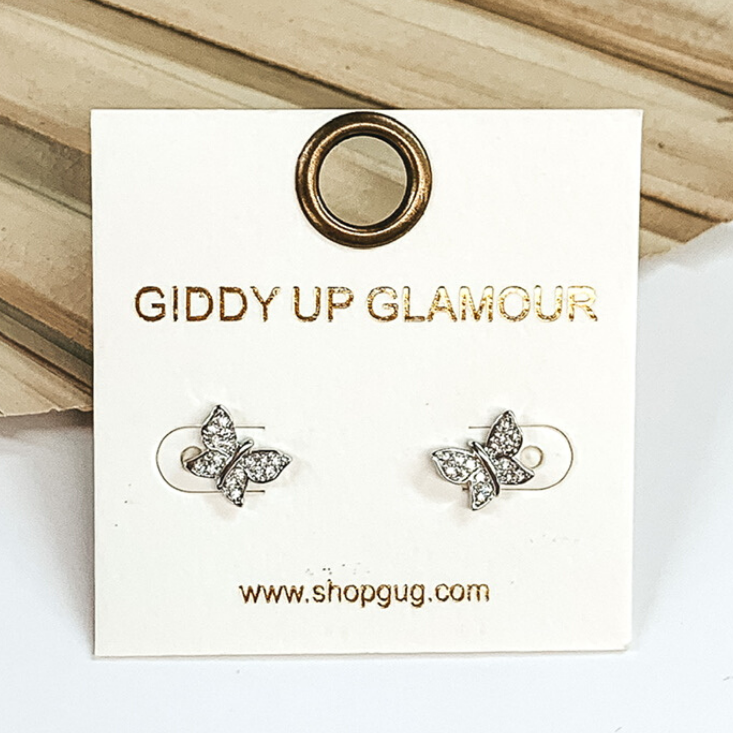 Small, silver butterfly stud earrings with clear crystals. These earrings are pictured on a white earrings card on a white and brown background.