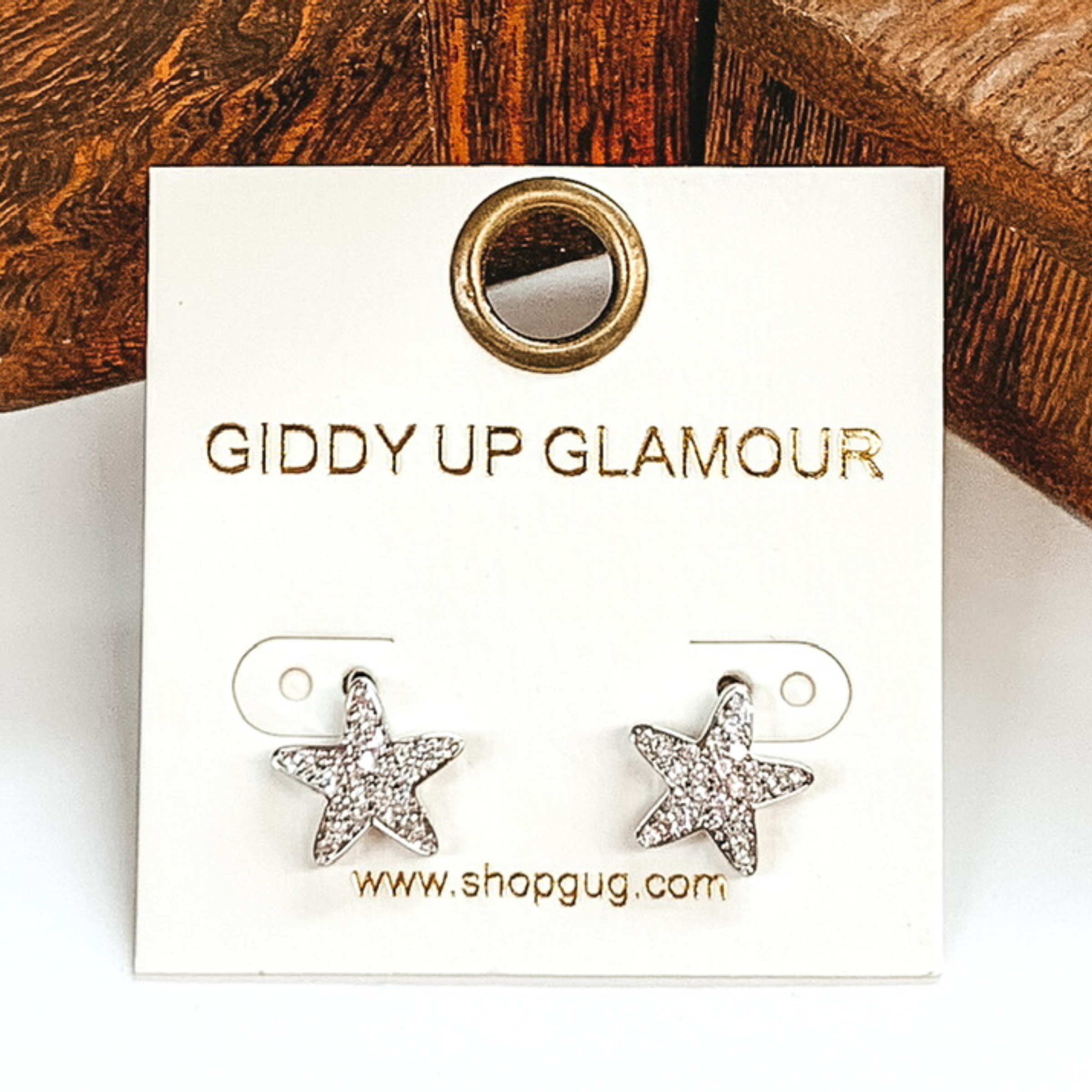 Small, silver star shaped stud earrings with clear crystals. These earrings are pictured on a white earrings card on a white and brown background.