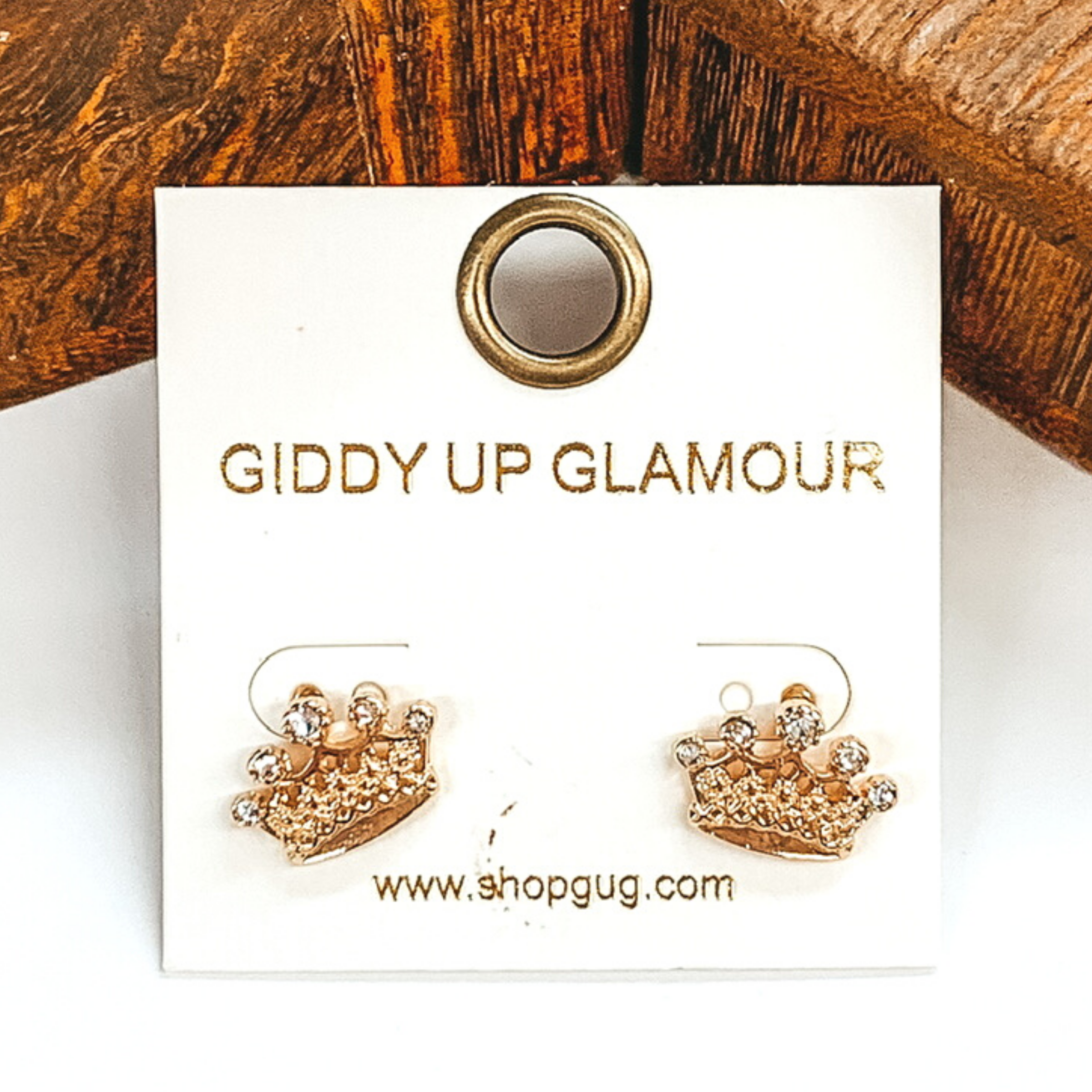 Gold crown stud earrings with clear crystals. These earrings are pictured on a white and brown background.