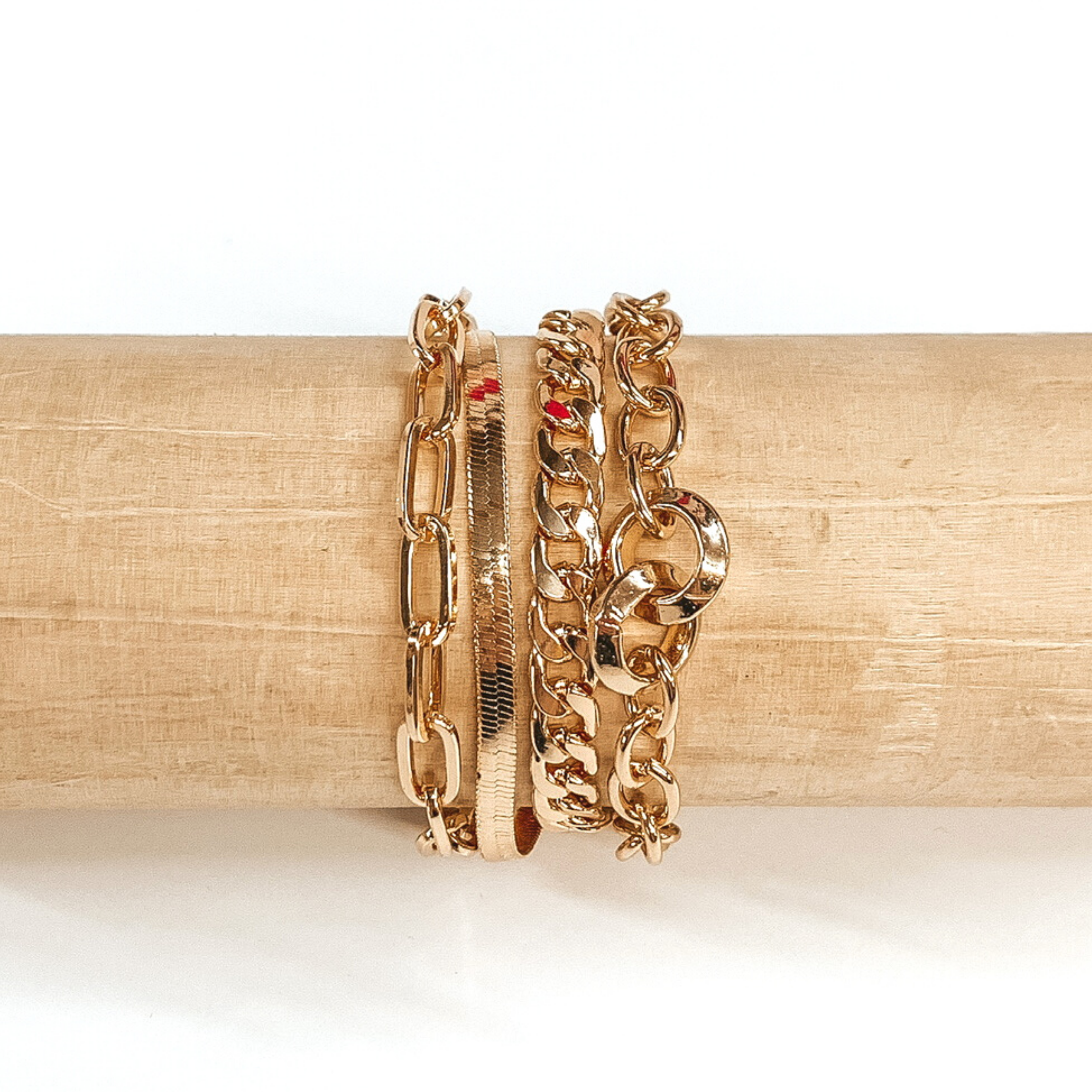 Four stranded gold bracelet pictured on a wood bracelet holder on a white background. This bracelet includes a paperclip chain, a curb chain, and a snacke chain, and a circle chain with two bigger chain  links. 