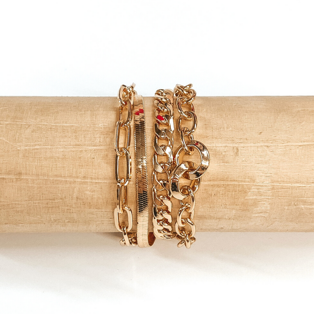 Four stranded gold bracelet pictured on a wood bracelet holder on a white background. This bracelet includes a paperclip chain, a curb chain, and a snacke chain, and a circle chain with two bigger chain  links. 