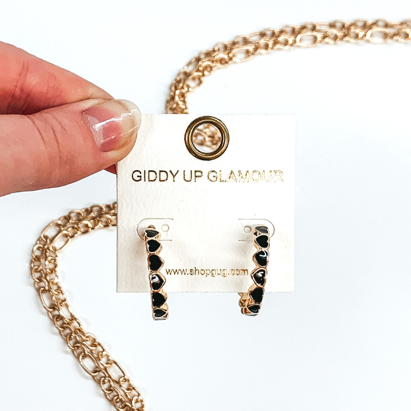 Gold outlined black heart that are connected to each other to form a hoop earrings. These hoops are pictured on a white earring holder held by fingers on a white background. 