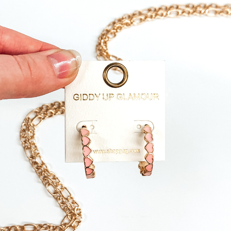 Gold outlined, light pink hearts that are connected to each other to form hoop earrings. These hoops are pictured on a white earring holder held by fingers on a white background with gold chains in the background.