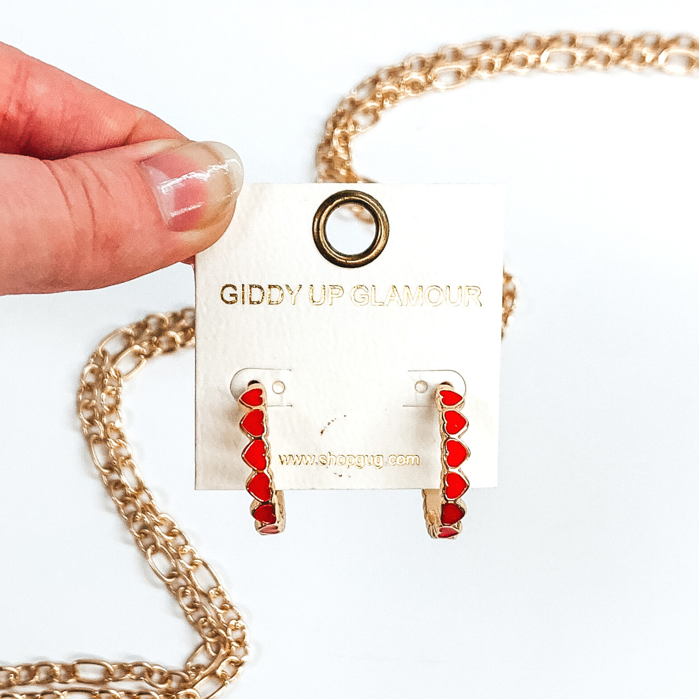 Gold outlined, red hearts that are connected to each other to form hoop earrings. These hoops are pictured on a white earring holder held by fingers on a white background with gold chains in the background.