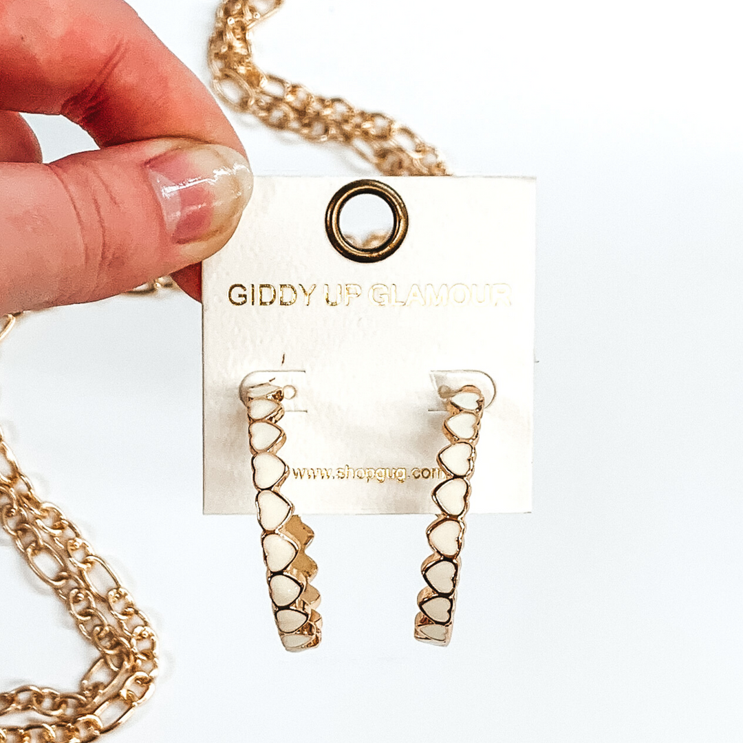 Gold outlined, white hearts that are connected to each other to form hoop earrings. These hoops are pictured on a white earring holder held by fingers on a white background with gold chains in the background.