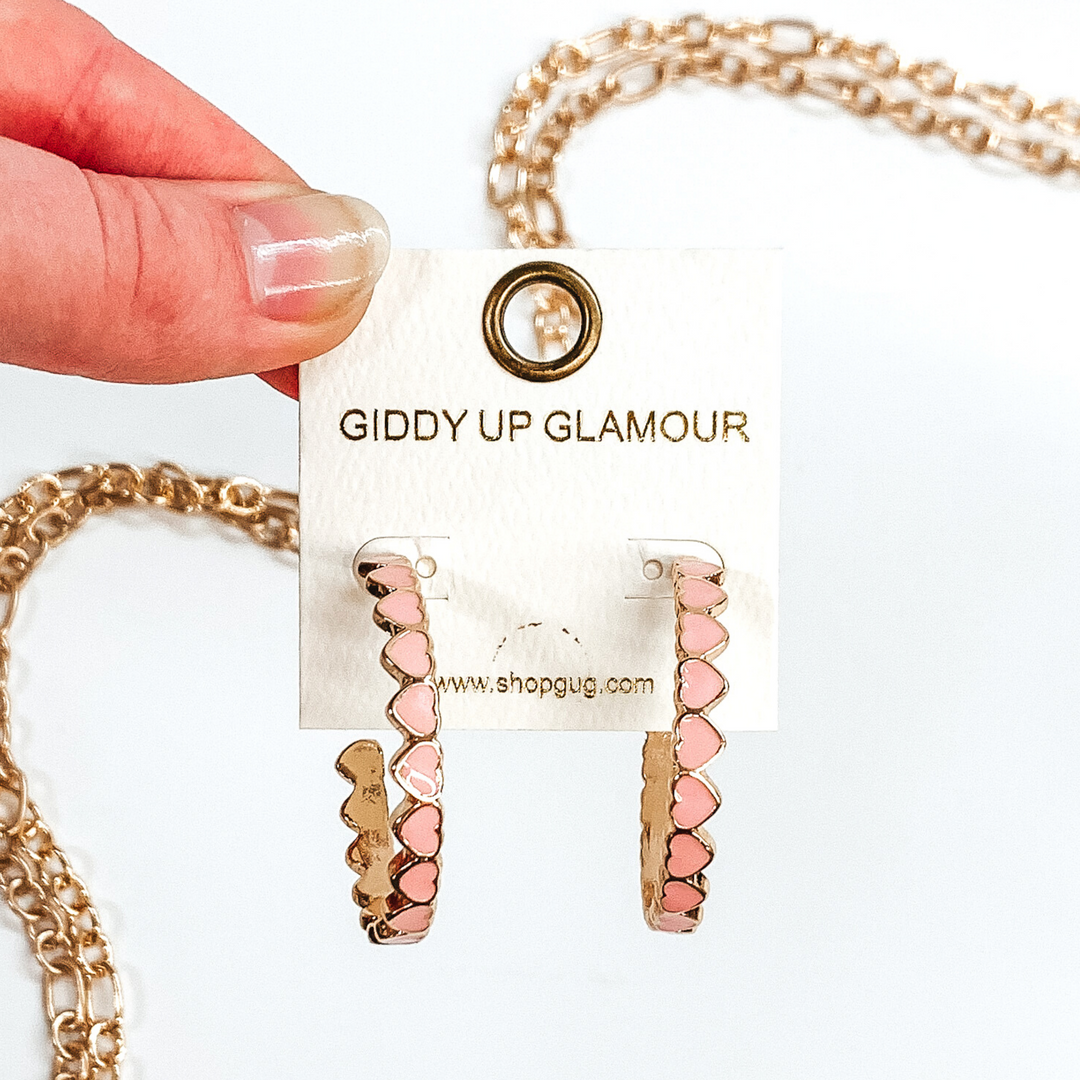 Gold outlined, light pink hearts that are connected to each other to form hoop earrings. These hoops are pictured on a white earring holder held by fingers on a white background with gold chains in the background.