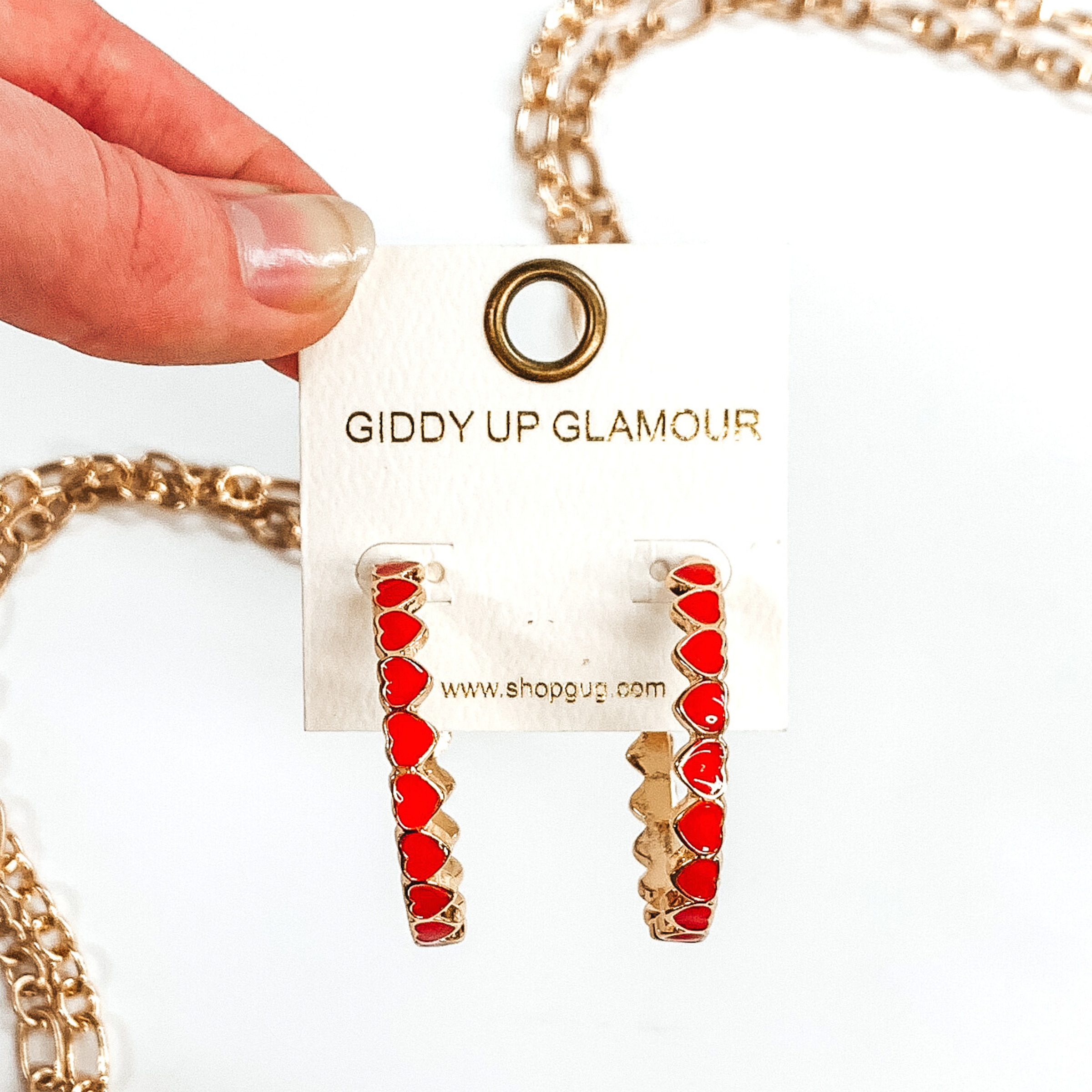 Gold outlined, red hearts that are connected to each other to form hoop earrings. These hoops are pictured on a white earring holder held by fingers on a white background with gold chains in the background.