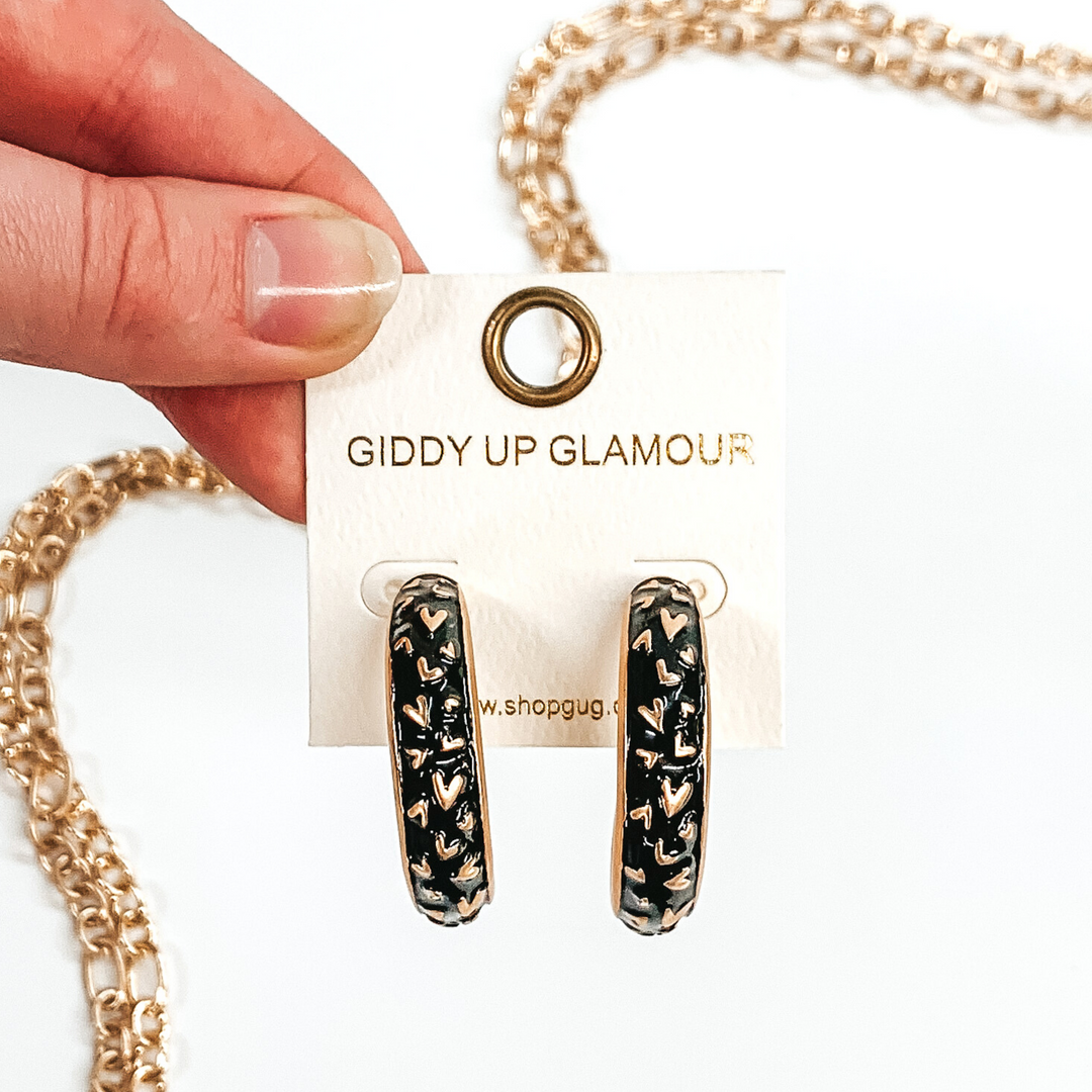 Thick, rounded black hoop earrings with gold outlined and gold heart pattern. These earrings are on a white earring holder held by fingers on a white background that has gold chained decor.