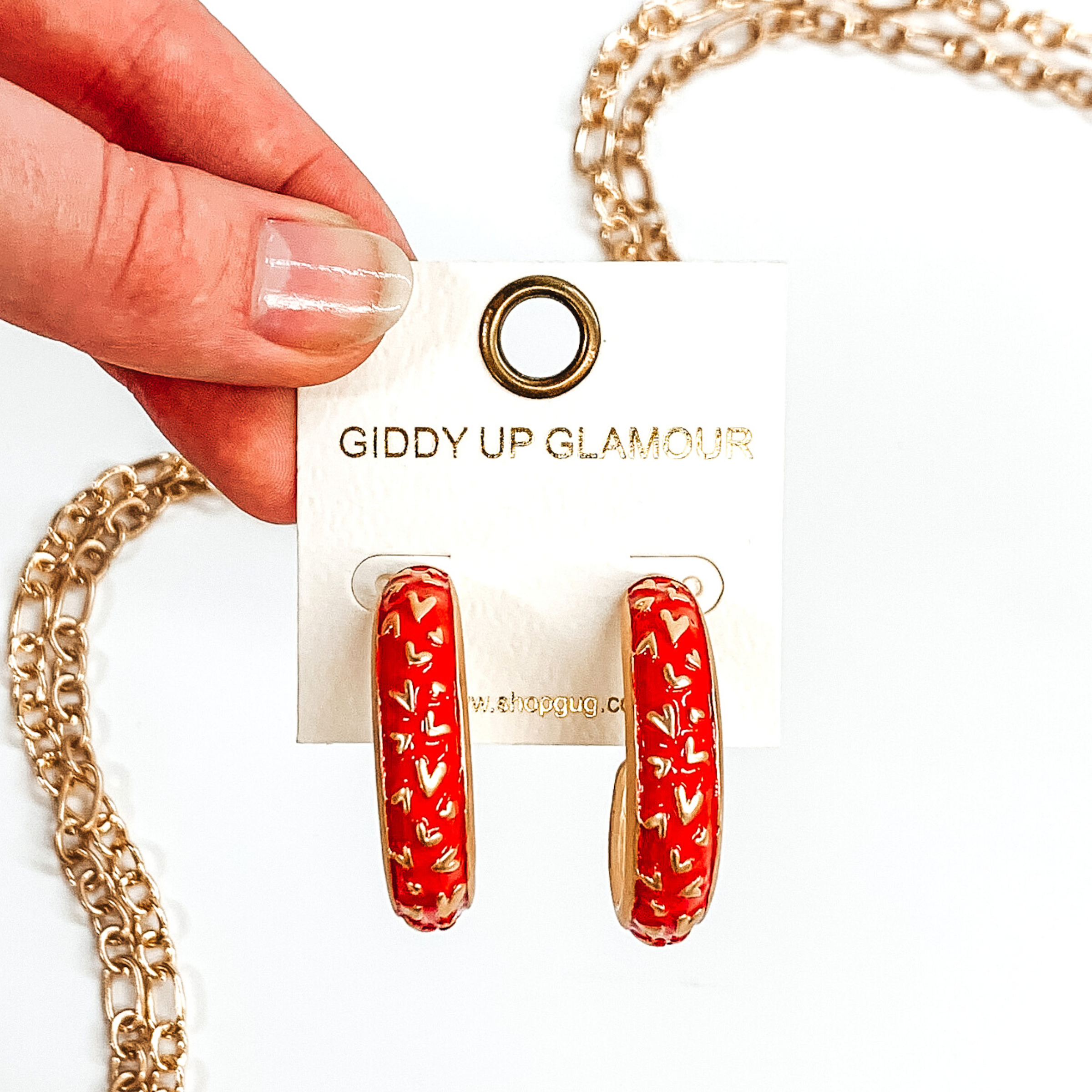 Thick, rounded red hoop earrings with gold outlined and gold heart pattern. These earrings are on a white earring holder held by fingers on a white background that has gold chained decor.