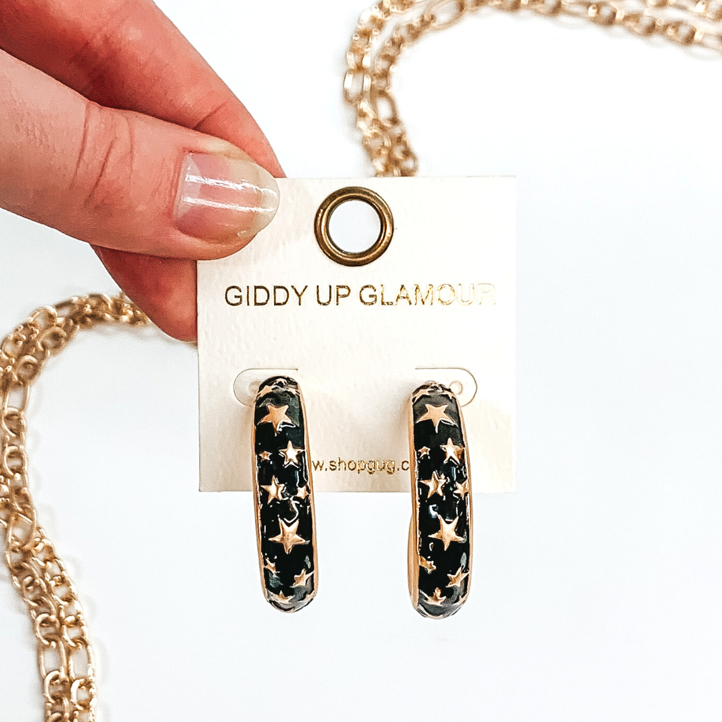 Thick, rounded black hoop earrings with gold outlined and gold star pattern. These earrings are on a white earring holder held by fingers on a white background that has gold chained decor. 