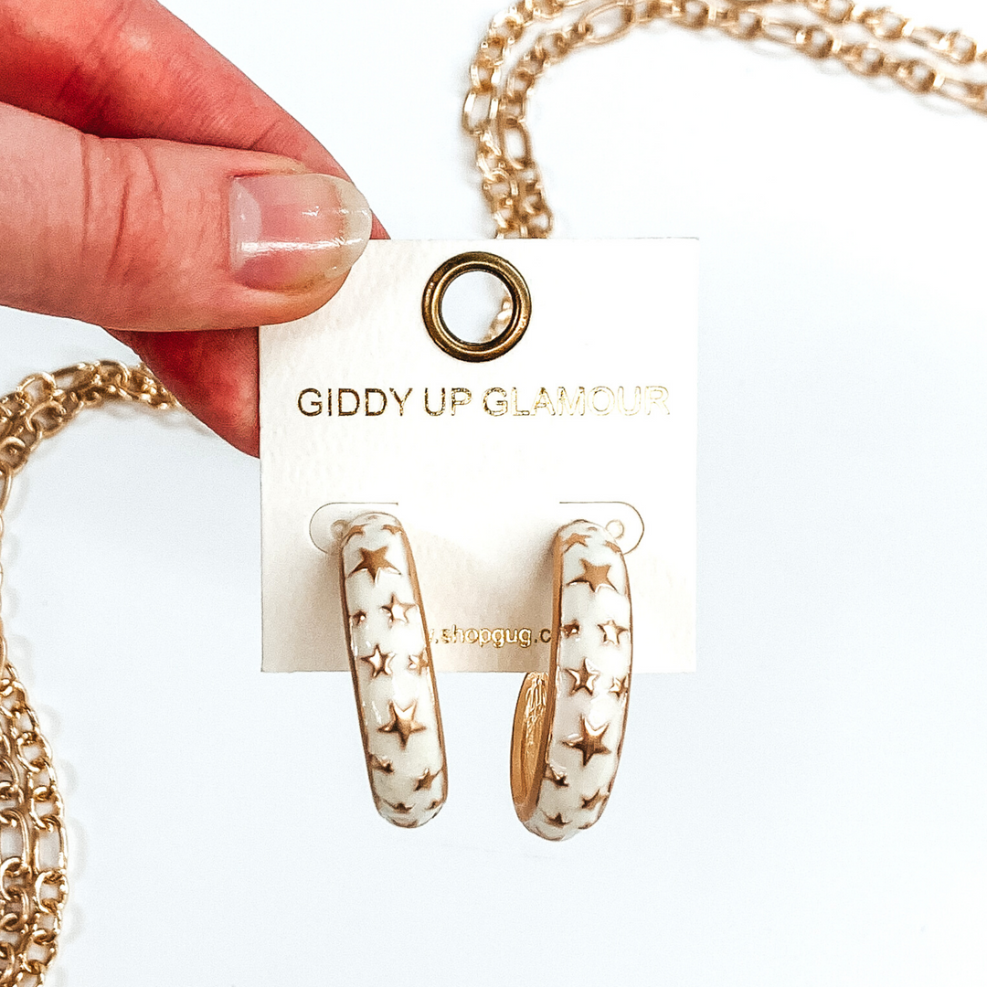 Thick, rounded ivory hoop earrings with gold outlined and gold star pattern. These earrings are on a white earring holder held by fingers on a white background that has gold chained decor.