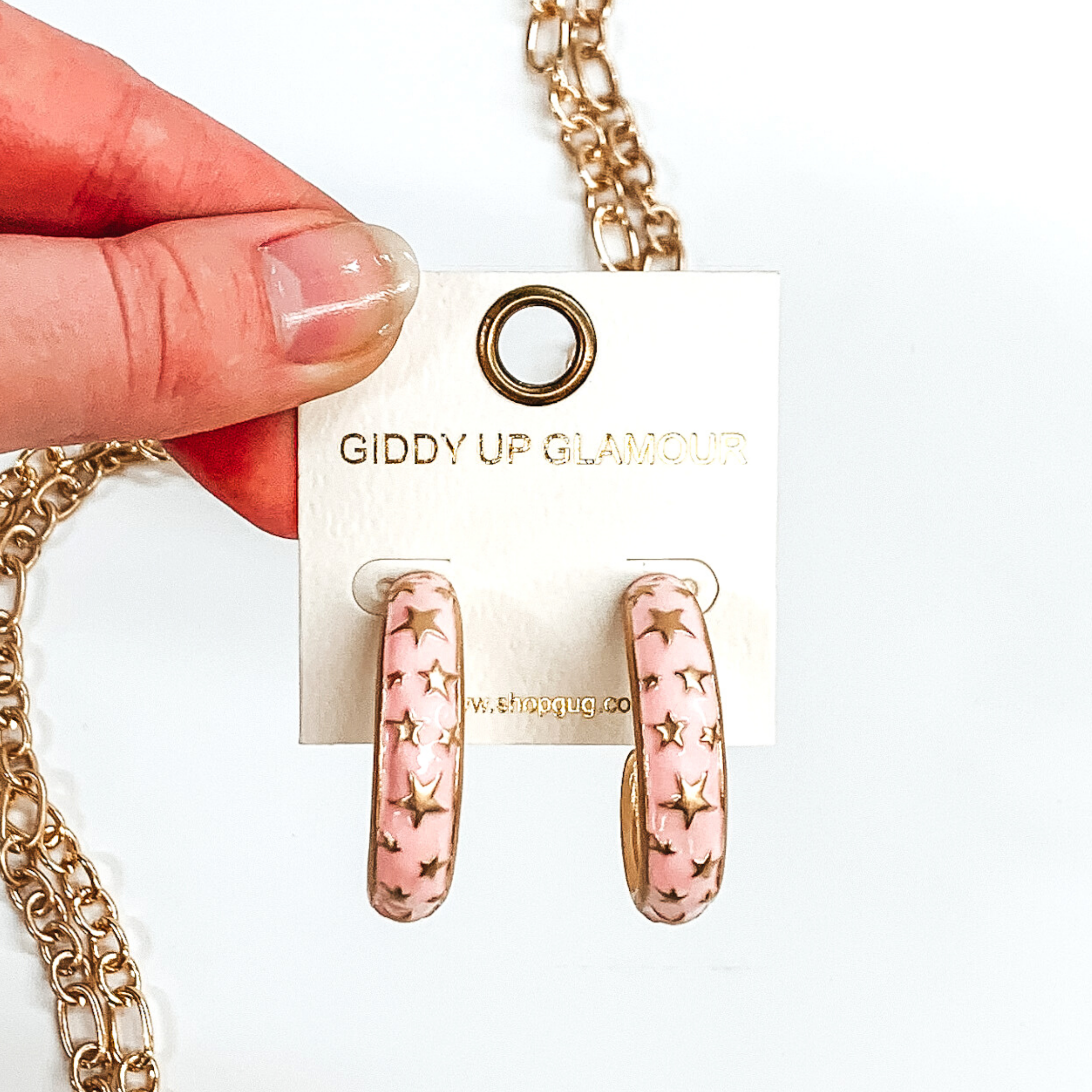 Thick, rounded light pink hoop earrings with gold outlined and gold star pattern. These earrings are on a white earring holder held by fingers on a white background that has gold chained decor. 