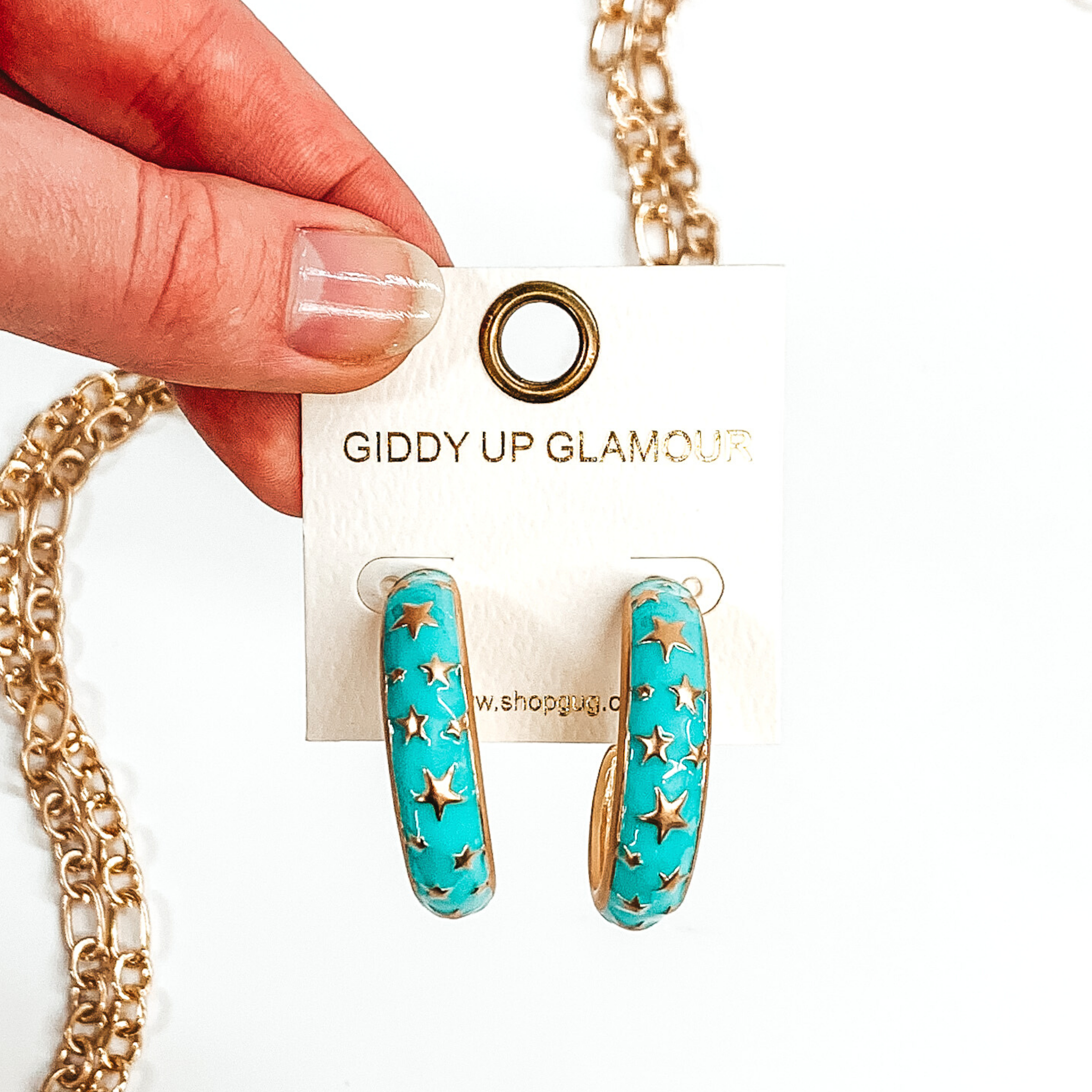 Thick, rounded light blue hoop earrings with gold outlined and gold star pattern. These earrings are on a white earring holder held by fingers on a white background that has gold chained decor. 