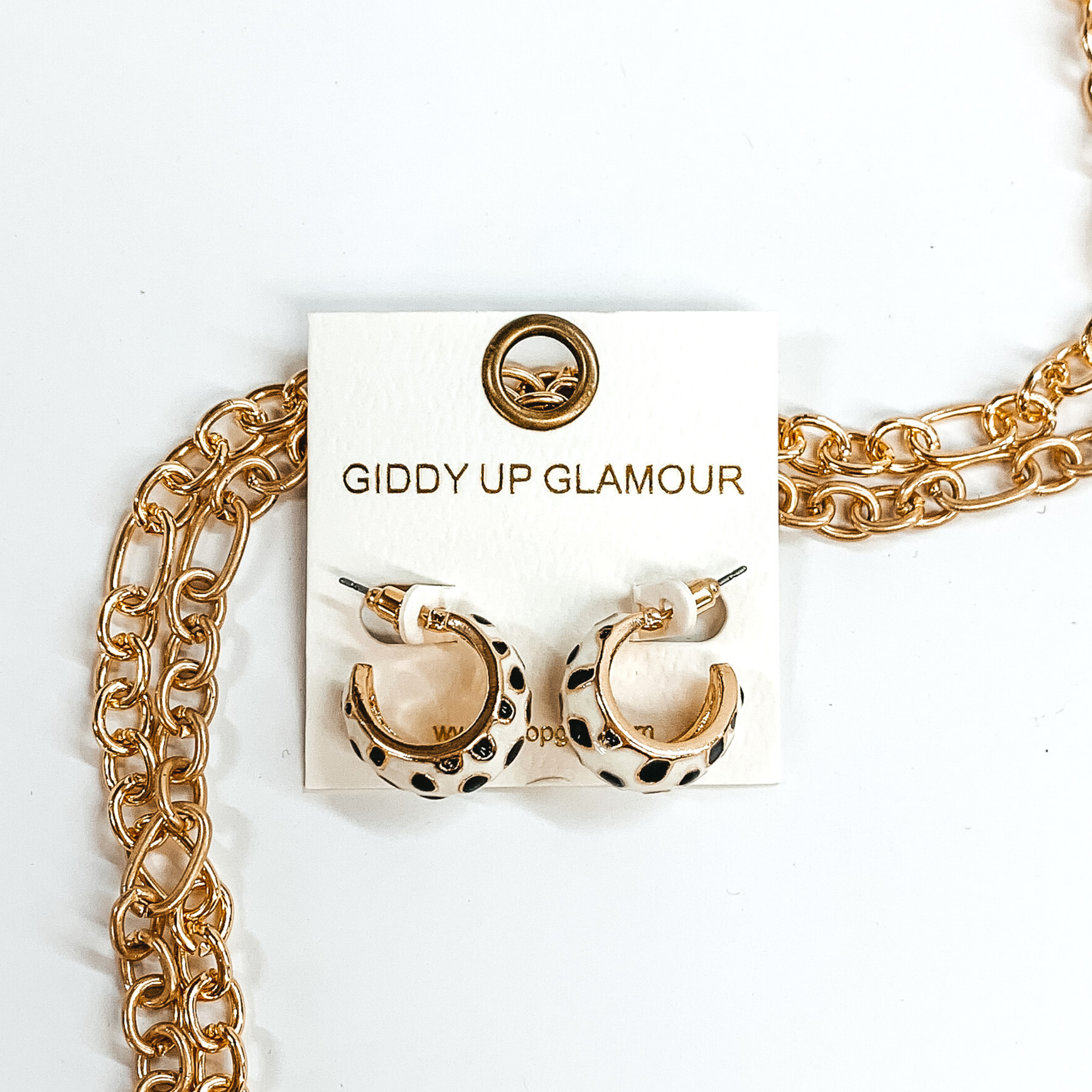 Mini, gold outline, white hoop huggies with black cheetah print.These earrings are pictured on a white earrings holder, laying on top of gold chains on a white background.