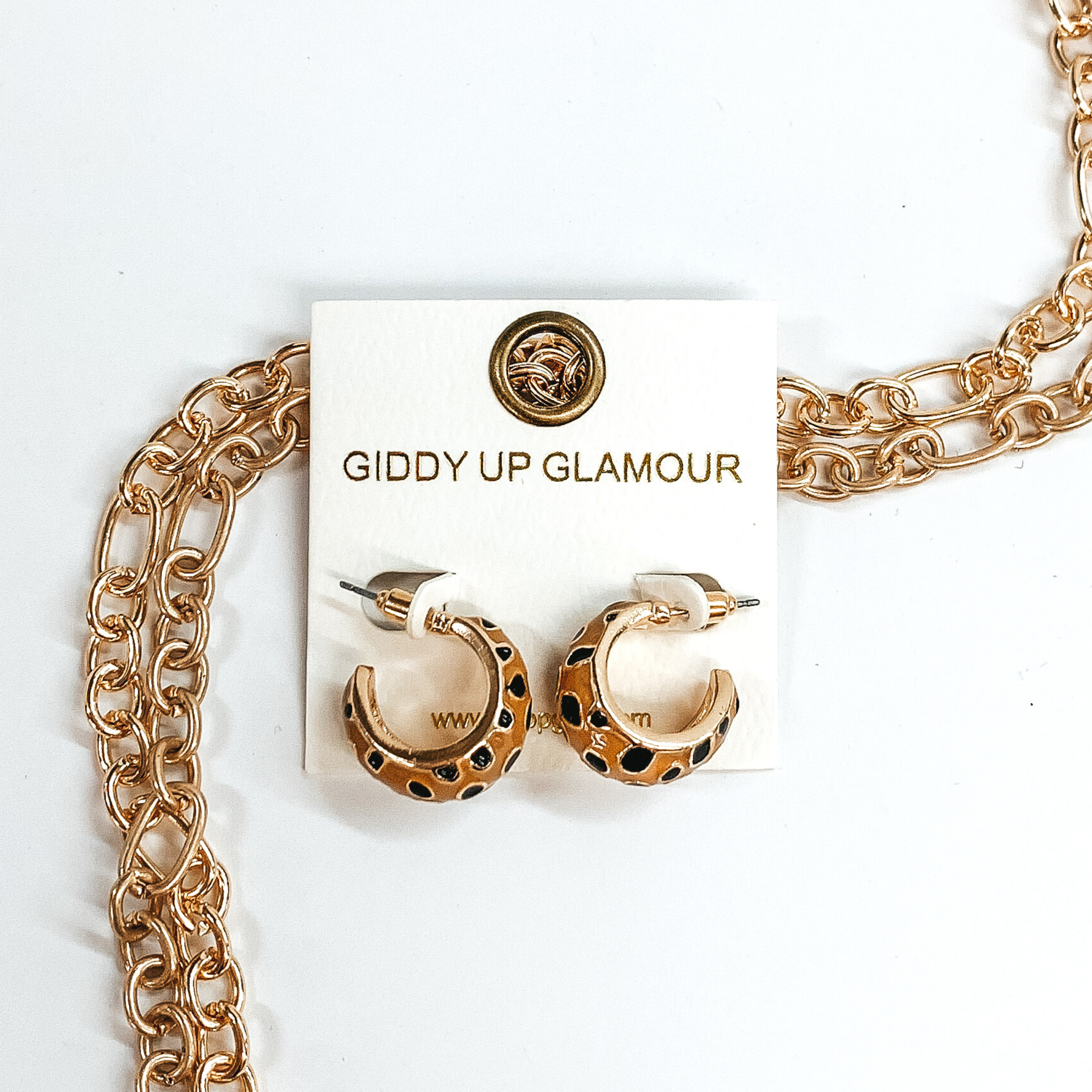 Mini, gold outline, tan hoop huggies with black cheetah print.These earrings are pictured on a white earrings holder, laying on top of gold chains on a white background. 