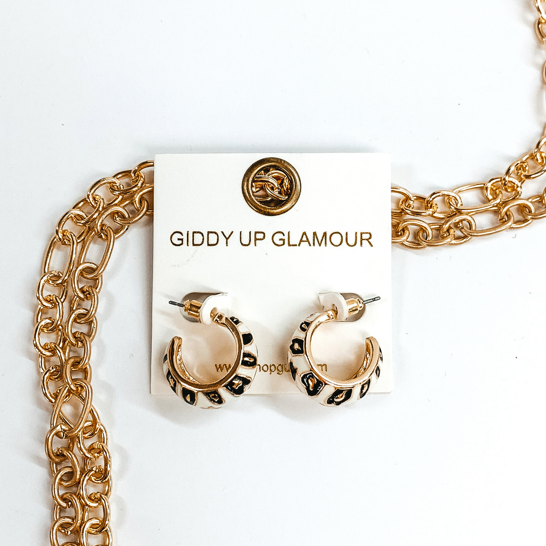 Mini, gold outline, white hoop huggies with black and gold leopard print.These earrings are pictured on a white earrings holder, laying on top of gold chains on a white background.