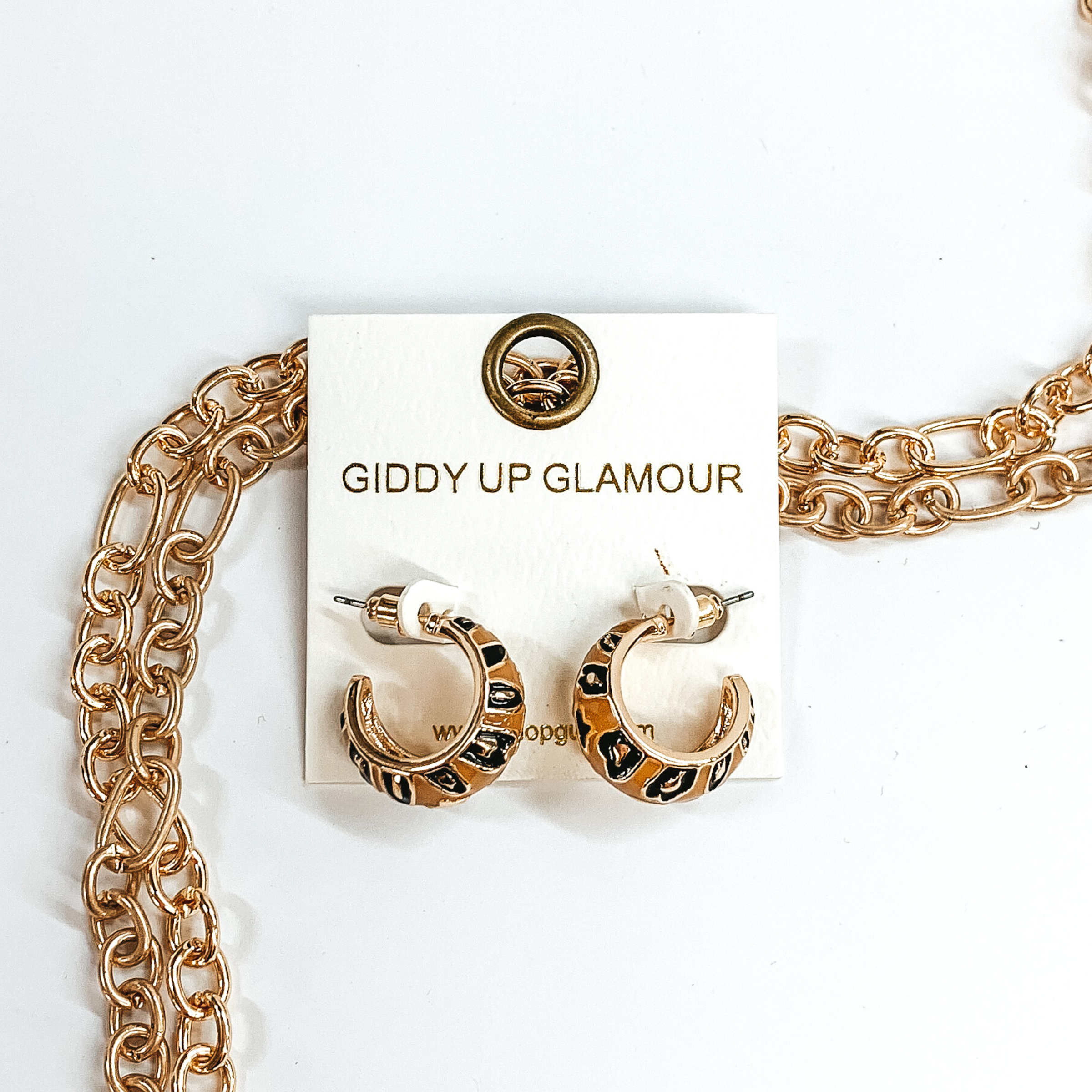 Mini, gold outline, tan hoop huggies with black and gold leopard print.These earrings are pictured on a white earrings holder, laying on top of gold chains on a white background.