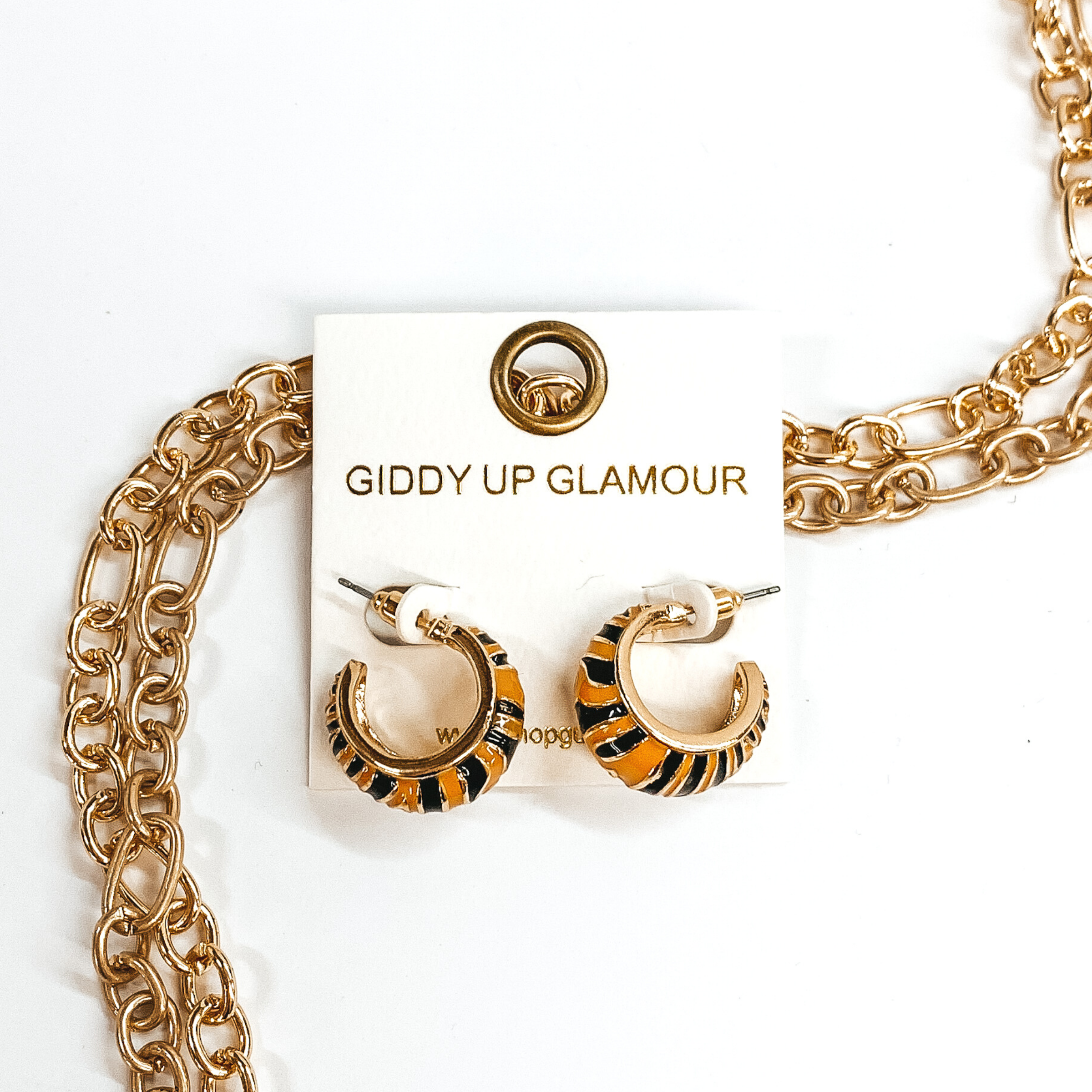 Mini, gold outline, tan hoop huggies with black tiger print.These earrings are pictured on a white earrings holder, laying on top of gold chains on a white background.