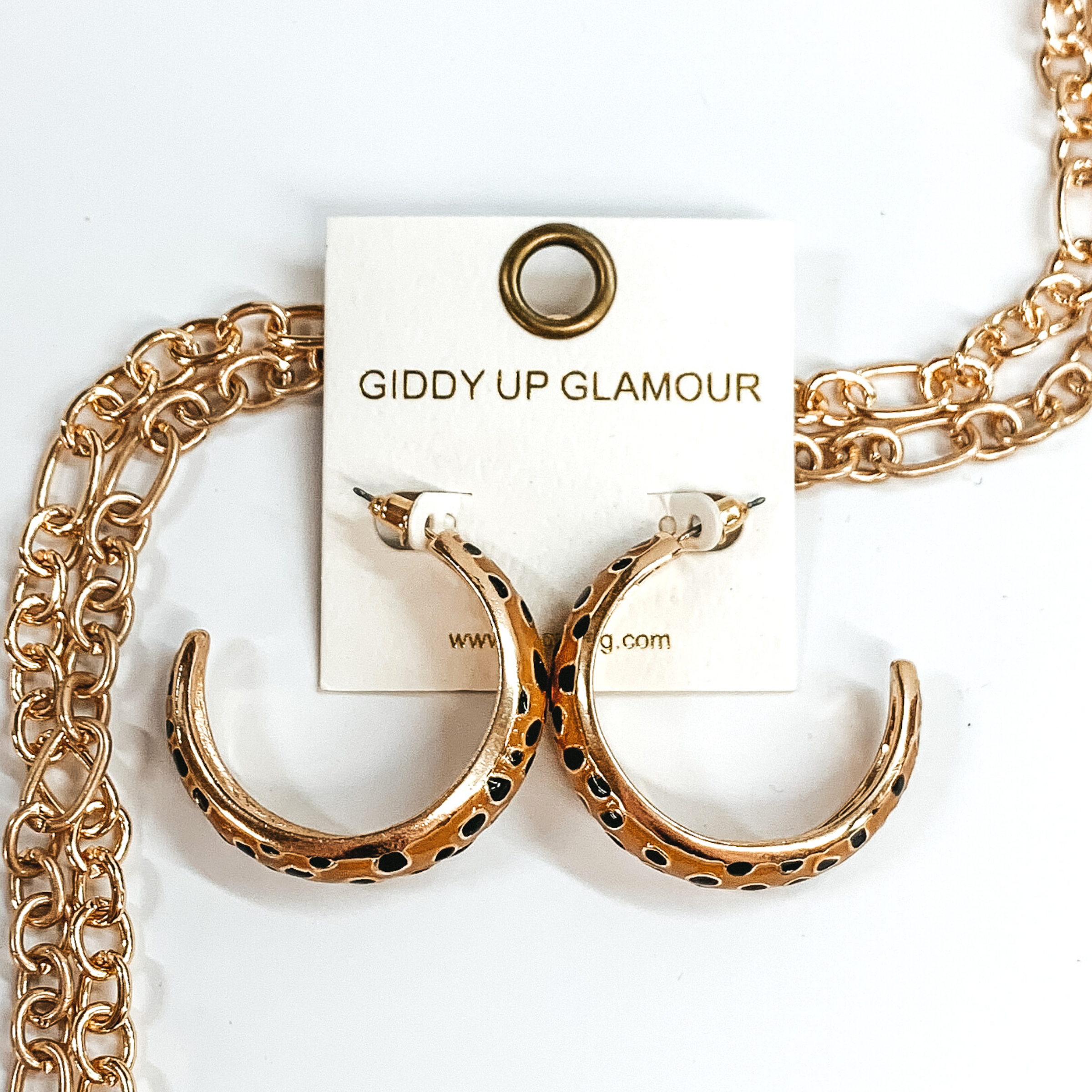 Large, gold outline, tan hoop earrings with black cheetah print.These earrings are pictured on a white earrings holder, laying on top of gold chains on a white background.