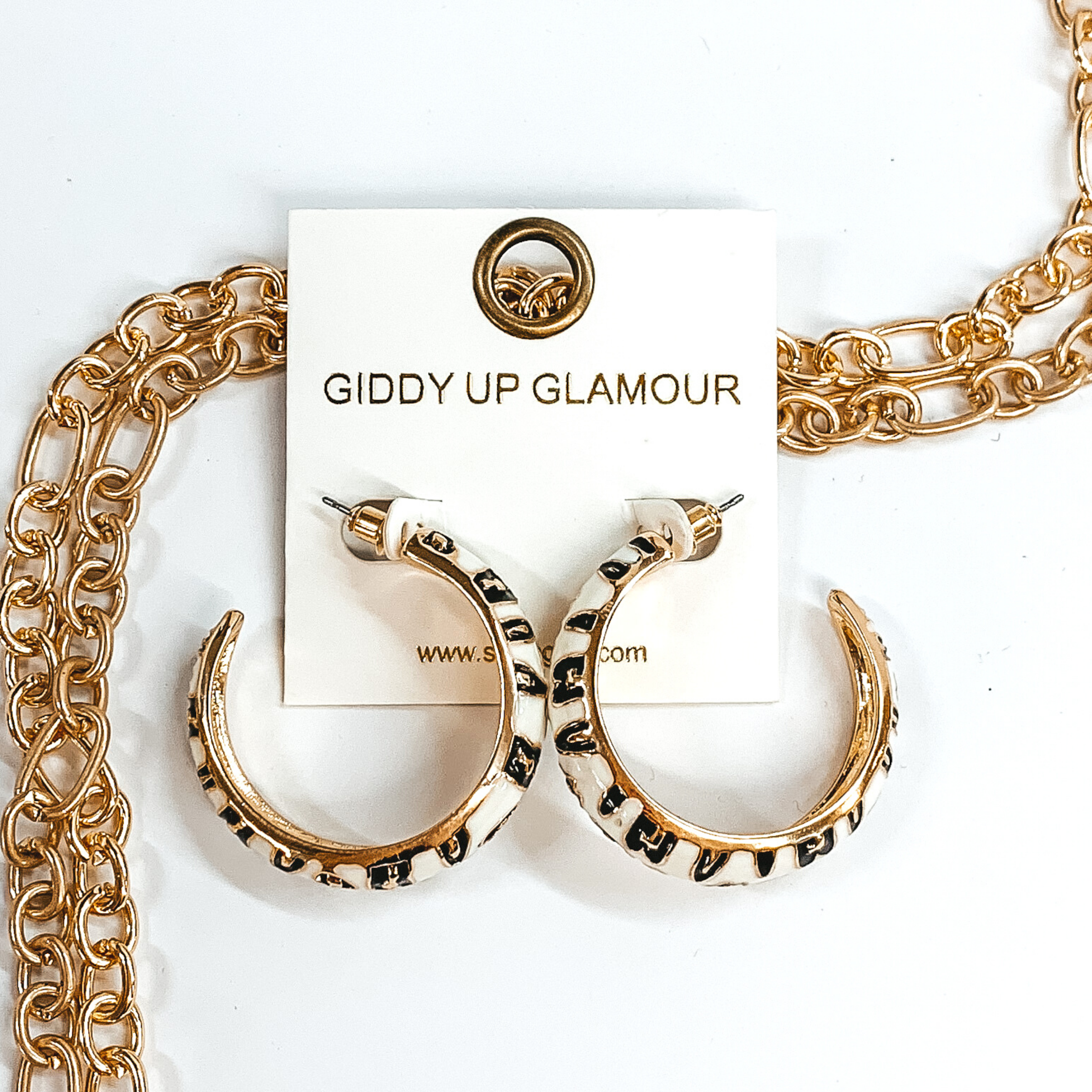 Large, gold outline, white hoop earrings with black and gold leopard print.These earrings are pictured on a white earrings holder, laying on top of gold chains on a white background.
