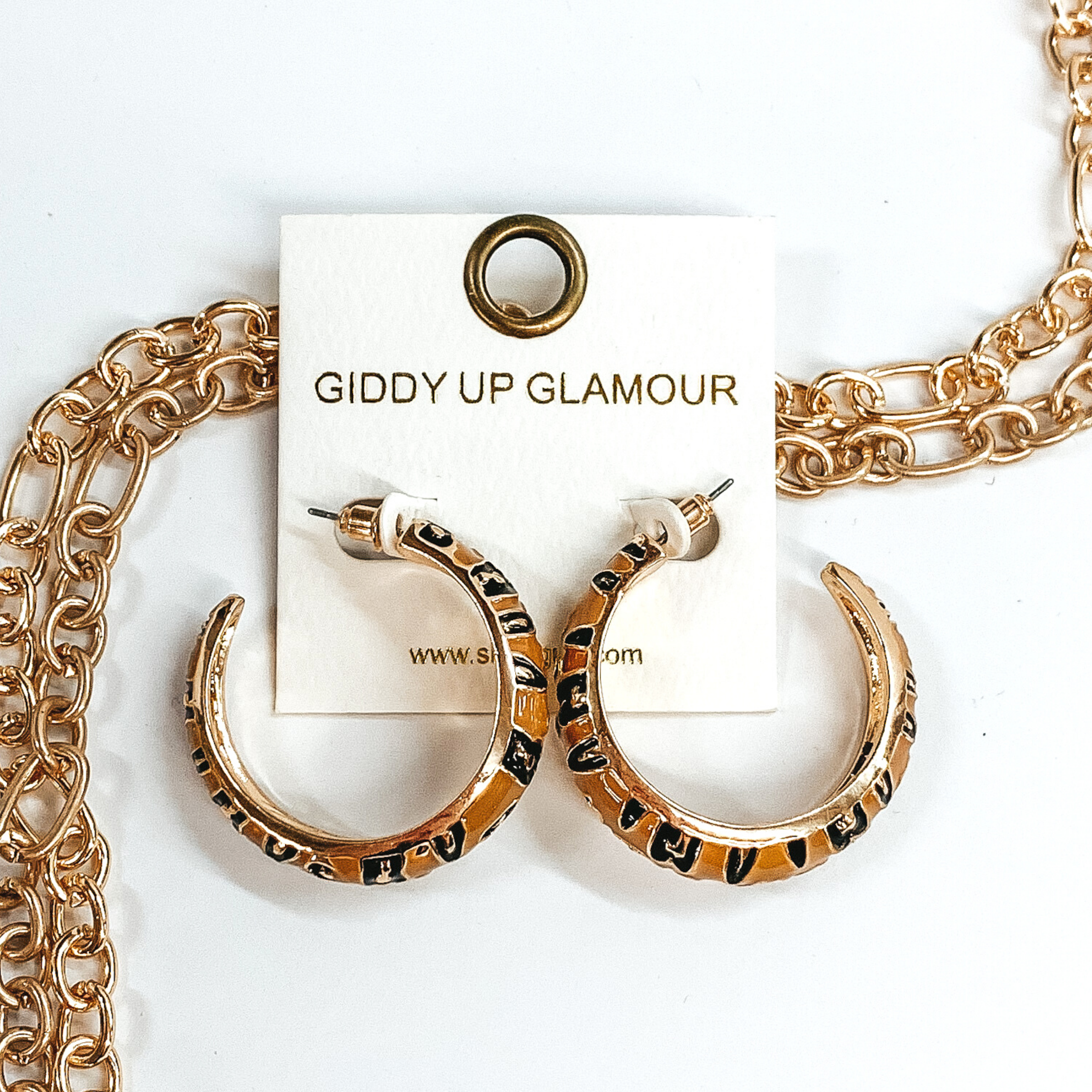 Large, gold outline, tan hoop earrings with black and gold leopard print.These earrings are pictured on a white earrings holder, laying on top of gold chains on a white background. 