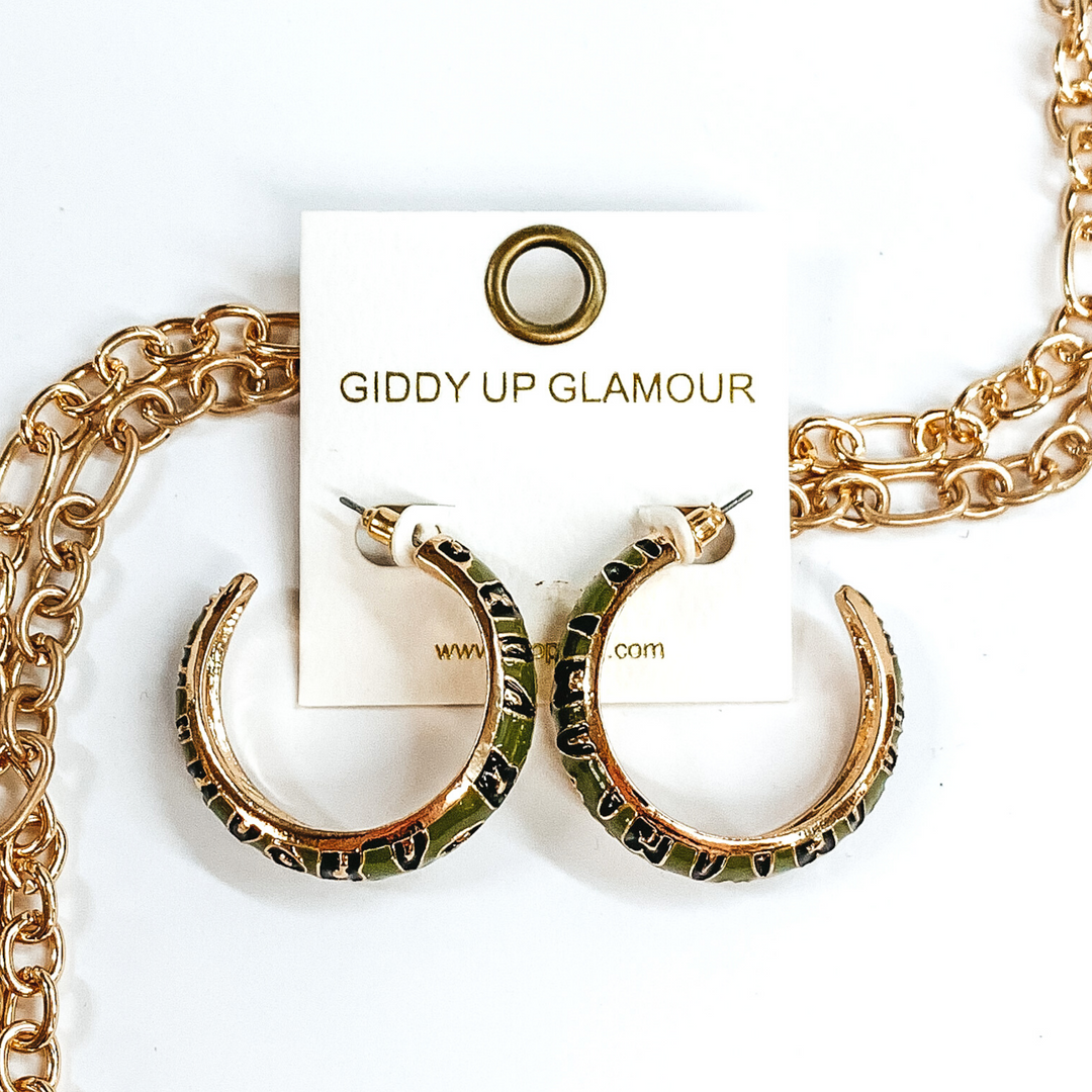 Large, gold outline, olive green hoop earrings with black and gold leopard print.These earrings are pictured on a white earrings holder, laying on top of gold chains on a white background.