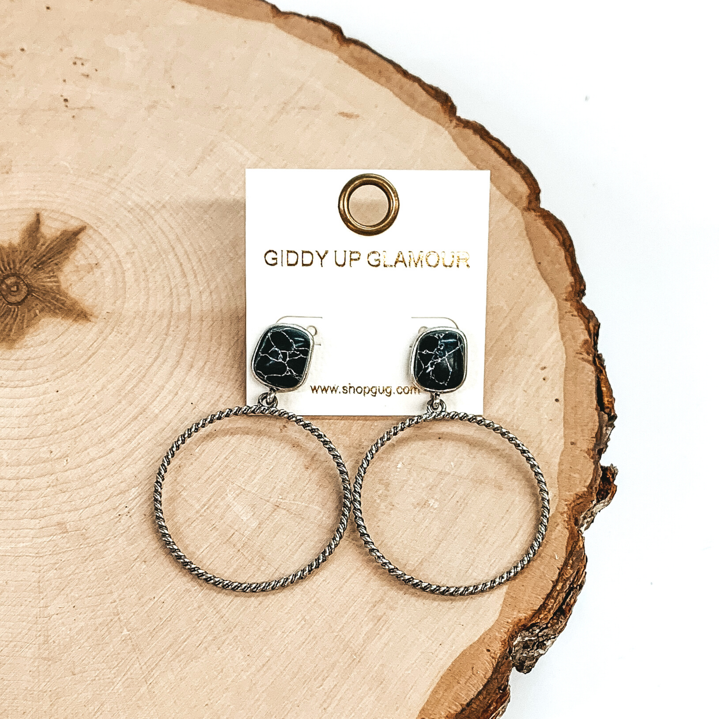 Rounded, square stone stud earrings in black with a silver, twisted open circle pendant hanging from the bottom. These earrings are pictured on a piece of wood on a white background.